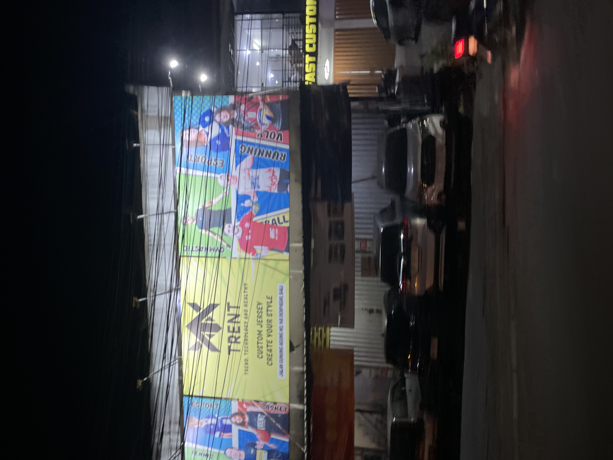 The image shows a night scene in Denpasar, Indonesia, focusing on a large billboard advertisement and the street in front of it. The billboard, illuminated by lights, advertises "TRENT TREND, TECHNOLOGY AND HEALTHY CUSTOM JERSEY CREATE YOUR STYLE" with images of people in athletic wear. Below the billboard, there is a storefront with signage that reads "FAST CUSTOM". Several cars are parked on the street in front of the building, and a motorcycle with its brake lights on is visible on the right side of the frame. The street appears to be paved, and there are some overhead wires and poles visible in the upper portion of the image. The overall impression is a commercial area at night, with a focus on custom apparel services.