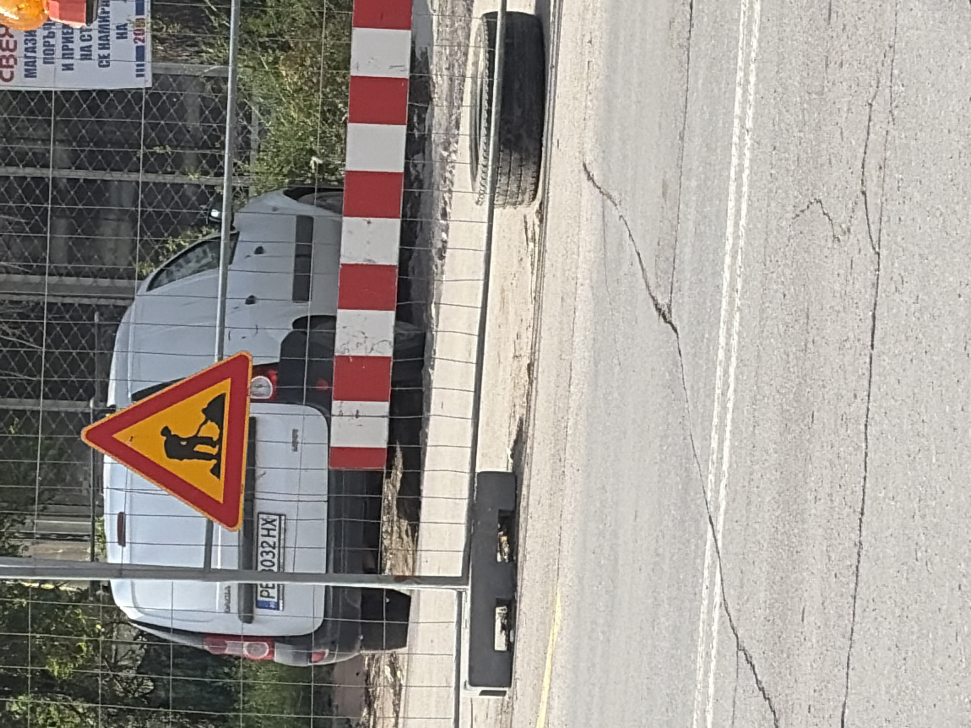 The image depicts a daytime roadside scene, likely a construction or maintenance area, viewed through a metal chain-link fence. A prominent red and white striped barrier pole stands vertically, partially obscuring the view.
Behind the fence, a white Dacia van is parked, facing away from the viewer. A yellow triangular warning sign with a red border, featuring a black silhouette of a worker shoveling (commonly known as a "men at work" sign), is placed in front of the van's rear. The van's Bulgarian license plate, "PE 032 HX," is clearly visible. Further behind the fence and to the left, a large white sign with Cyrillic text is partially visible, containing words such as "МАГАЗИН" (store), "ПОРЪЧ..." (order), and "СЕ НАМИРА" (is located), indicating an advertisement or informational board. Green foliage is visible in the background through the fence.
To the right of the fence and barrier is an asphalt road surface. A single white lane marking runs along the road, which shows numerous cracks. A dark car tire leans against the curb between the fence and the road. The bright lighting suggests it is a clear, sunny day. The Cyrillic text and the Bulgarian license plate confirm the location in Bulgaria, consistent with Plovdiv.