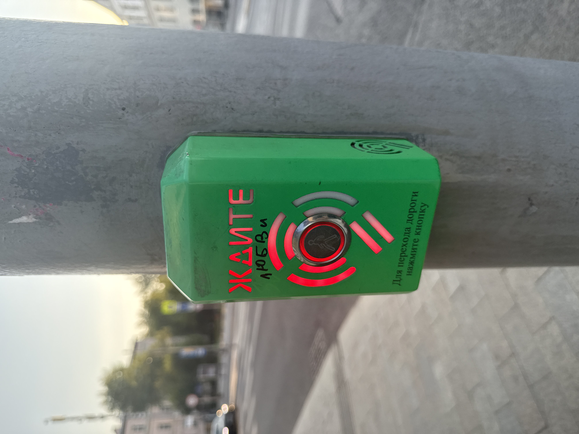 This media file shows a close-up of a green pedestrian crossing button attached to a metal pole. The button is illuminated with red lights, indicating an active signal. The text "ЖАЙТЕ ЛЮБВИ" (meaning "Wait for Love" or "Wait for Kindness" in Russian, likely a playful or artistic addition to a standard sign) is visible on the left side of the button in red letters. To the right, in smaller green text, it says "Для перехода дороги нажмите кнопку" (meaning "For crossing the road, press the button"). The button has a circular central area with a pictogram of a walking person, also illuminated in red, surrounded by concentric red arcs.
The background is blurred but suggests an outdoor urban environment, likely a street in Moscow, Russia, given the Cyrillic text. The time of day appears to be late afternoon or early evening, with soft, diffused light. The sky is bright, suggesting clear or partly cloudy weather. The overall impression is a functional object integrated into a city landscape, with the illuminated button suggesting an imminent pedestrian crossing.