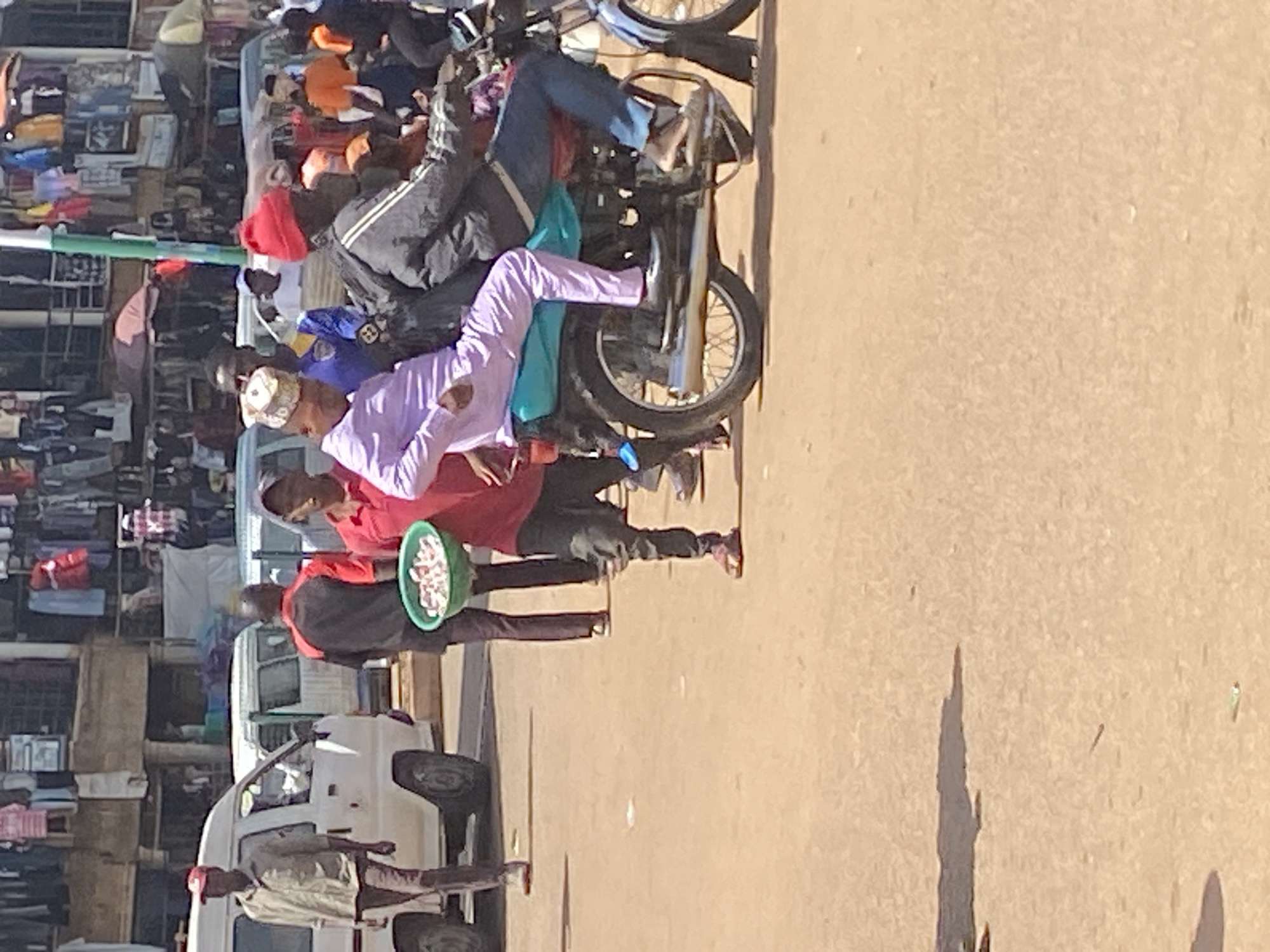 In Bukuru, Nigeria, a scene unfolds on a dusty street. Several people are present, some walking and others on a motorcycle. A man wearing a red cap and a light brown shirt walks near a white van. To his right, another man in a red and black shirt carries a green bowl filled with what appears to be food items. In the background, market stalls display various goods, including clothing. A motorcycle with two passengers rides past. One passenger, wearing a purple outfit and a patterned cap, is seated at the front. The second passenger, in a gray jacket and red hat, is behind him. The environment is typical of a bustling Nigerian market, with a mix of vehicles, pedestrians, and commercial activity under a bright sky.