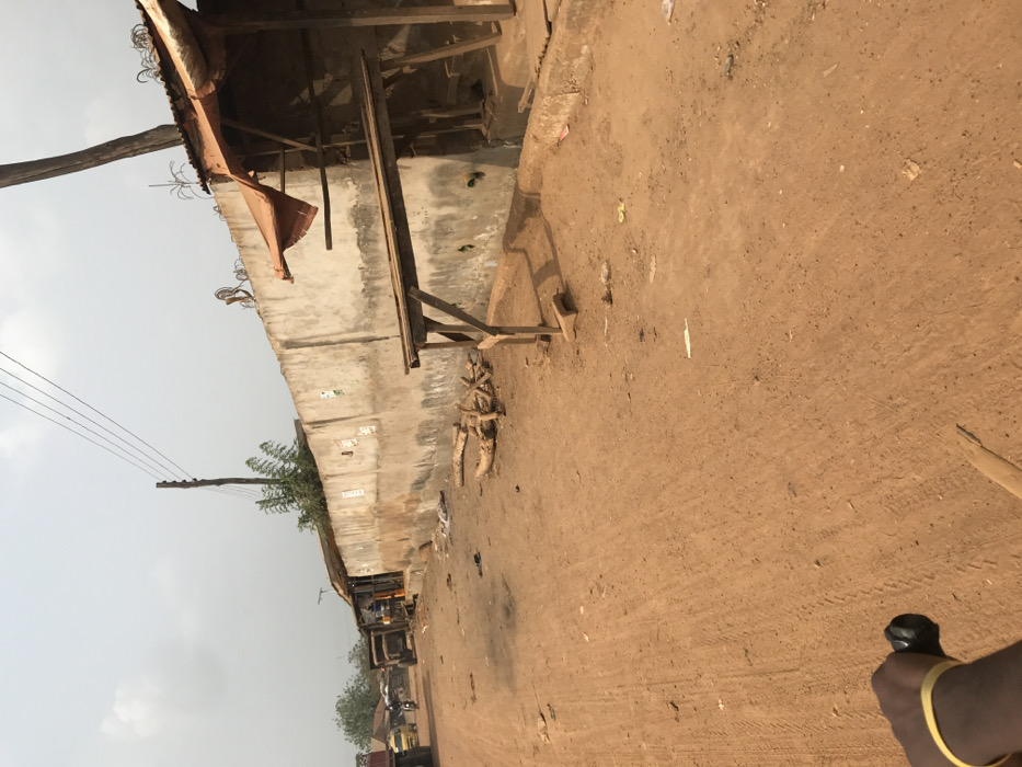 The image depicts an unpaved reddish-brown dirt road in Ogbese, Nigeria, under a hazy light-colored sky. The perspective is from the immediate foreground, with a partial view of a dark-skinned arm wearing a yellow wristband and holding a black handlebar-like object in the bottom-left corner.
Along the right side of the road, a long, weathered concrete wall extends into the mid-ground. Razor wire or barbed wire is affixed to the top of the wall in sections. Adjacent to the wall, a makeshift wooden stall with a corrugated metal roof covered by an orange tarp is visible. In front of this, a simple wooden table structure stands on the dirt. A pile of cut logs or firewood is located on the ground between these structures and the road. Scattered debris and litter are present on the ground.
On the left side of the road, utility poles with overhead power lines are visible. Further down the road, several structures with corrugated metal roofs line the street. Vehicles, including a yellow three-wheeled auto-rickshaw (Keke Napep) and a large dark truck, are parked or in motion. Multiple indistinct figures are visible near the vehicles and buildings in the mid-ground. Sparse green vegetation is visible behind the wall and structures in the distance.
