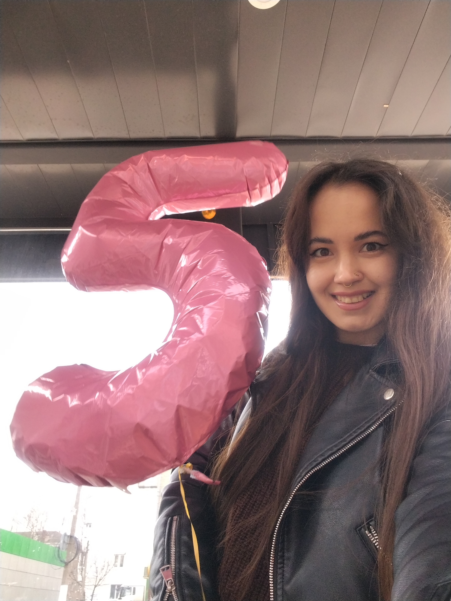 A smiling young woman holds a pink foil balloon shaped like the number 5. She wears a black leather jacket and stands beneath a dark, paneled awning or roof. The background shows a bright, overcast day with buildings and possibly trees visible in the distance. The setting appears to be Chaikovsky, Russia, based on contextual clues inferred from the visual elements.