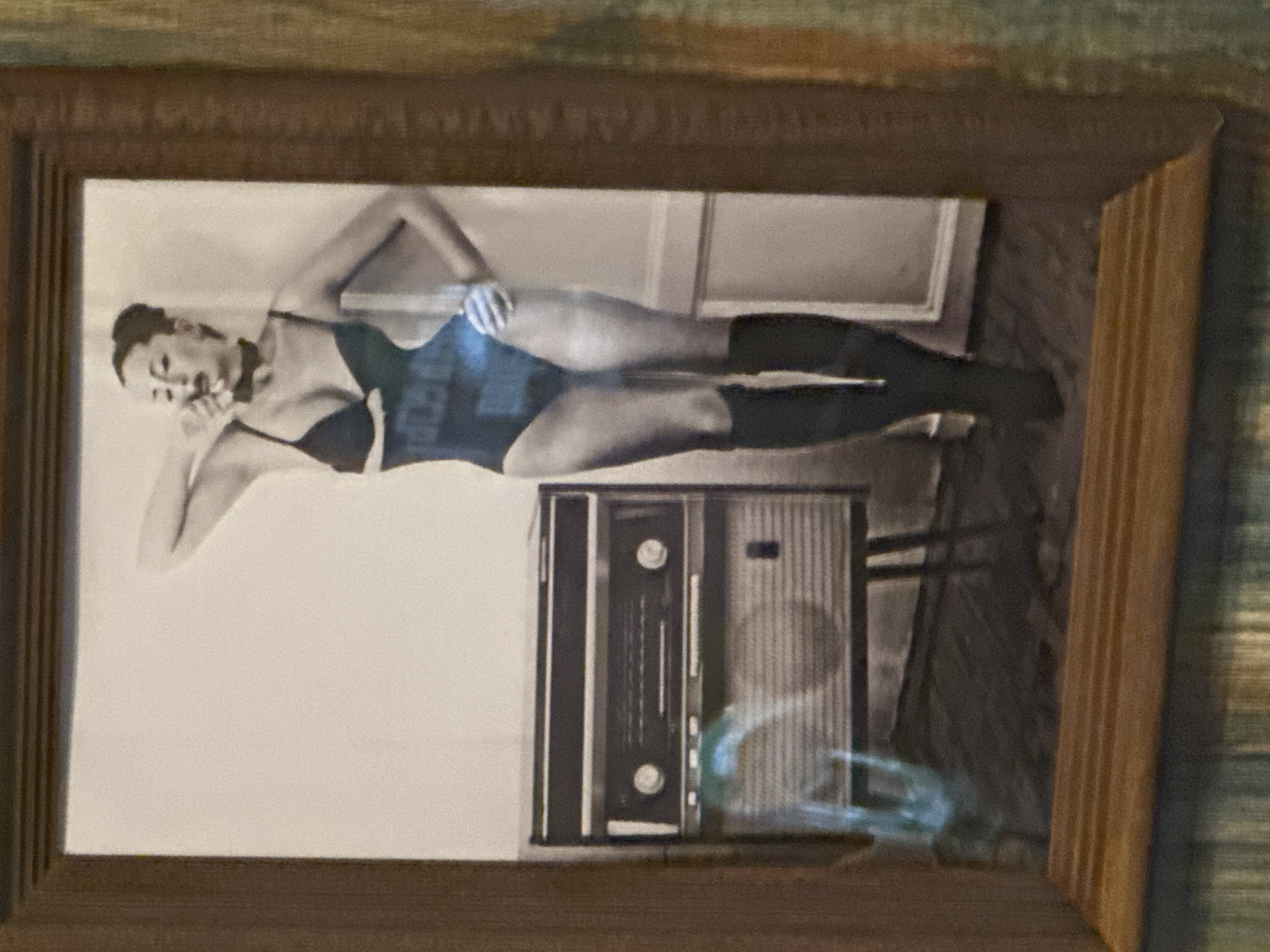 The image shows a framed black and white photograph. The photograph depicts a woman reclining in a one-piece bathing suit and knee-high boots next to an old radio. The location of this image is in Selyatino, Russia.