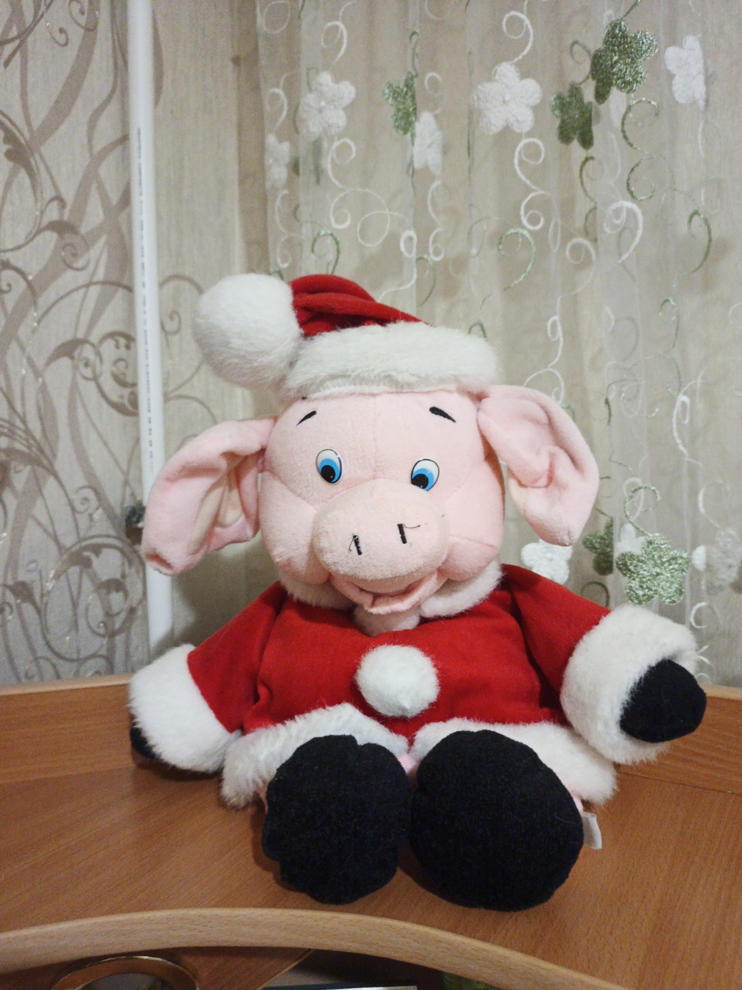 A plush pig toy wearing a Santa Claus hat and coat is sitting on a wooden surface. The pig is pink with blue eyes, a black nose, and black hooves. The Santa hat is red with a white trim and a white pom-pom. The coat is red with white trim and a white pom-pom on the chest. The background is a curtain with a floral pattern. The image is taken in a well-lit indoor environment.