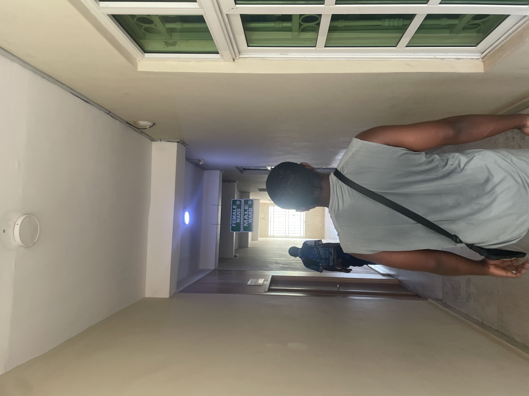 The image depicts a hallway in a building, with two individuals walking away from the camera. The hallway is lit by overhead lights, one of which emits a blue glow. Green signs indicating "FEMALE WARD" and "MALE WARD" are visible hanging from the ceiling, suggesting the location is likely a hospital or clinic. The walls are painted a light beige color, and a window with green-tinted glass is present on the right side of the hallway. The floor appears to be tiled. The individual in the foreground is wearing a light gray sleeveless shirt and a black bag slung across their shoulder. The person in the background is wearing a patterned shirt. The architectural style and the presence of signage suggest this could be a facility within the city of Abuja, Nigeria, possibly a hospital ward.