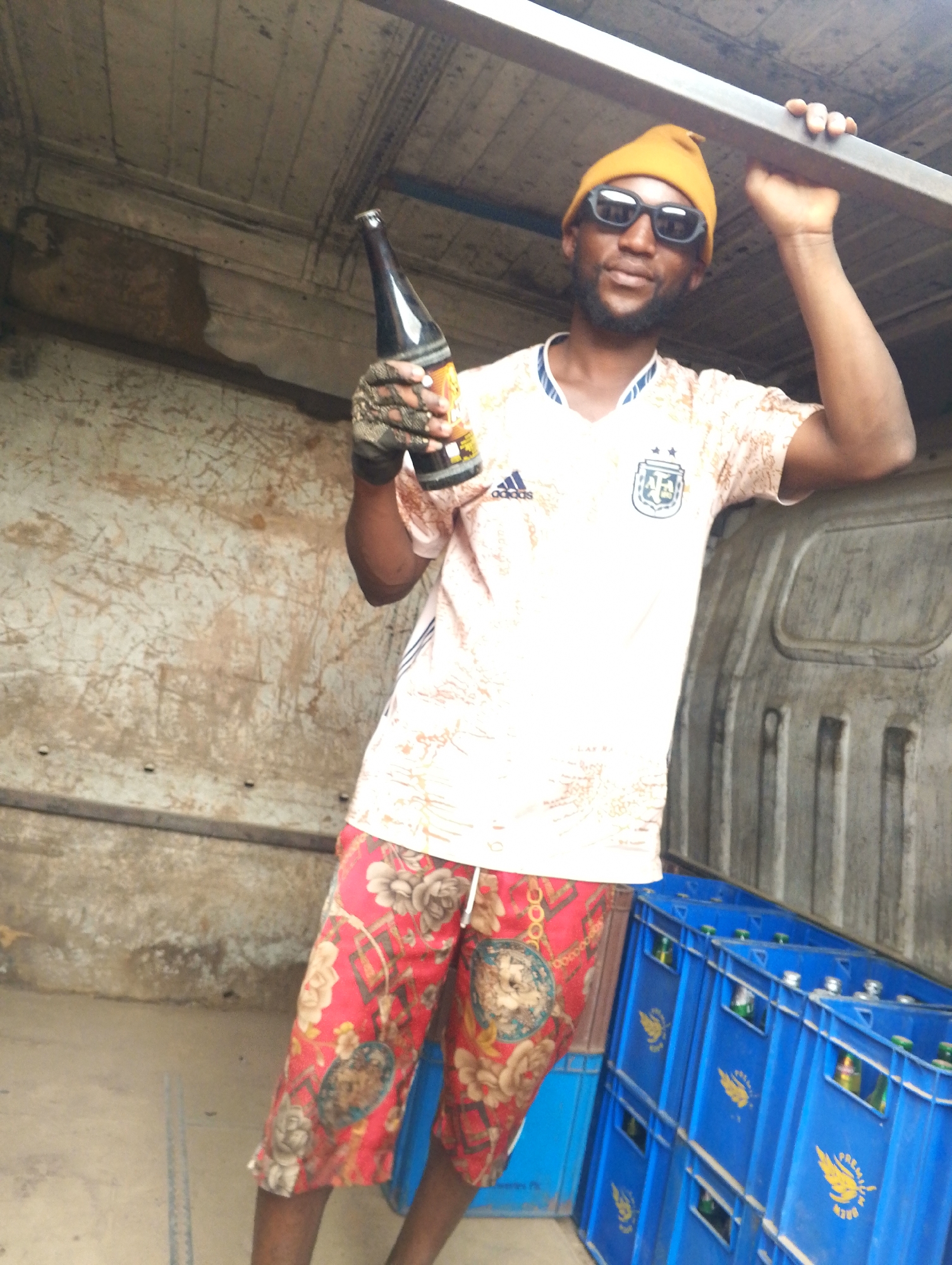 A man stands inside a vehicle, holding a bottle, in Unknown, Unknown. The man wears a yellow beanie, sunglasses, a white Argentina soccer jersey, and patterned red shorts. His right hand is covered by a gray glove. He holds a dark-colored bottle in his gloved hand. His left hand is gripping the metal frame of the vehicle. Behind him, blue crates containing bottles are visible. The interior of the vehicle shows signs of wear and is poorly lit.