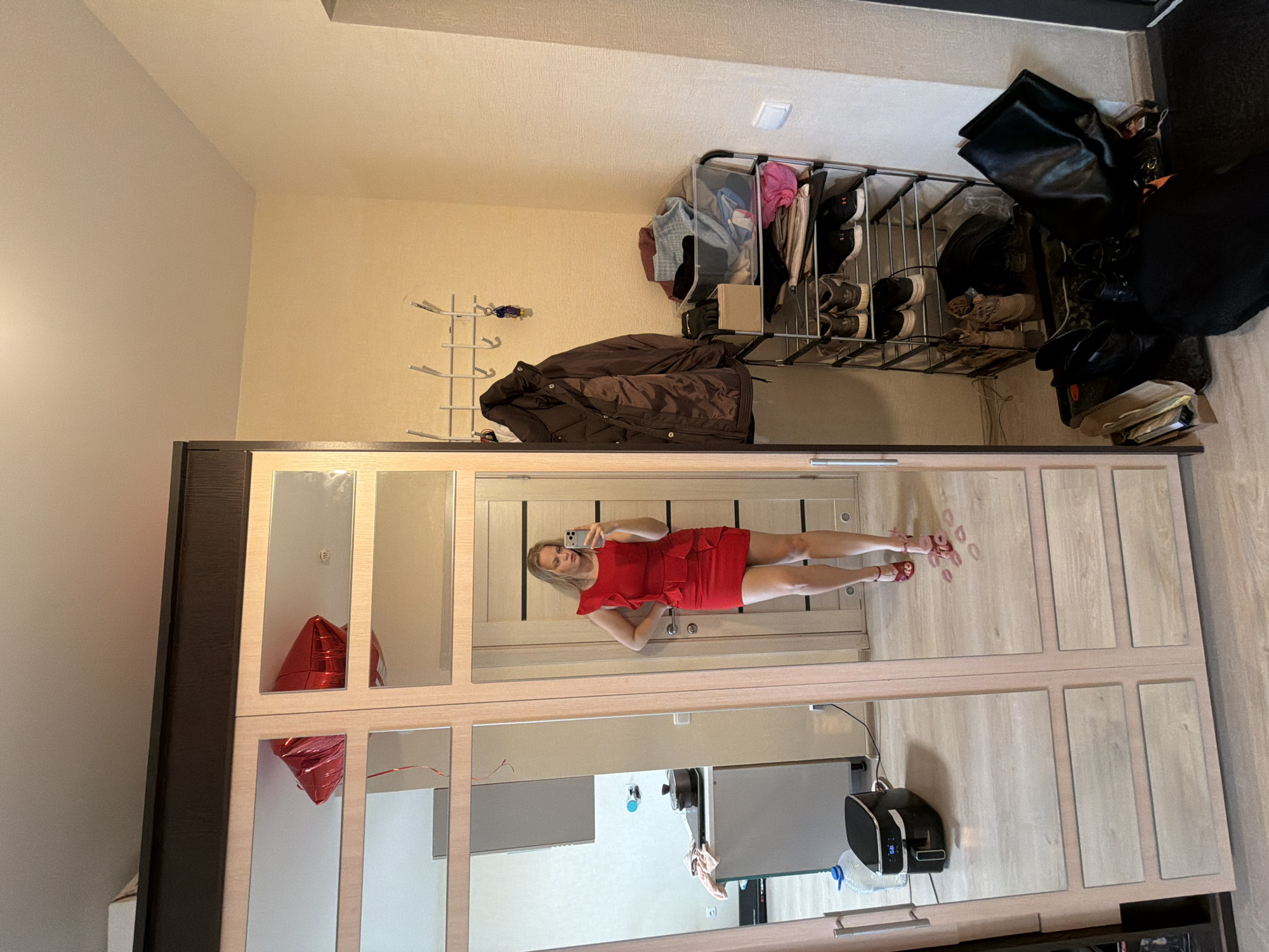 A person is reflected in a mirrored wardrobe. The person is a blonde woman wearing a short red dress and red sandals. She is holding a smartphone, presumably taking a picture of herself in the mirror. The surrounding environment includes a coat rack, a shoe rack with shoes and bags, and a red heart-shaped balloon inside the wardrobe. The image was taken in Izhevsk, Russia, as indicated by the file metadata.