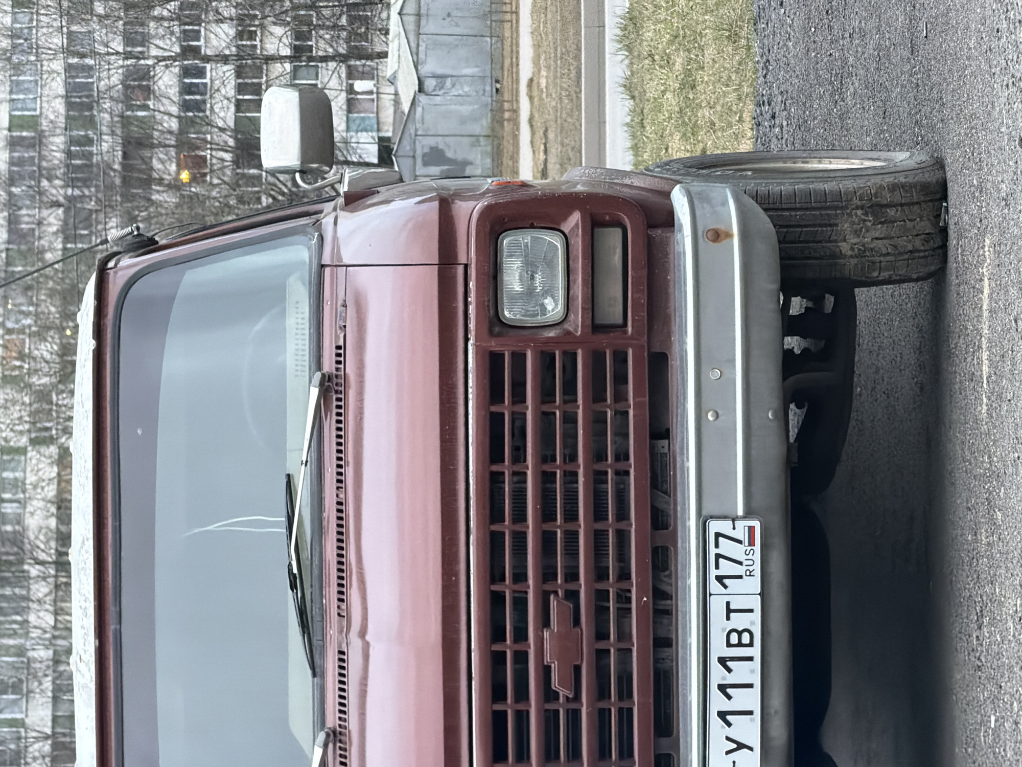 The image shows the front of a maroon Chevrolet van with a Russian license plate reading "У111ВТ 177 RUS". The van is parked on asphalt pavement. In the background, a building with many windows is visible, along with some greenery. Given the "177" license plate code, it indicates that the vehicle is likely registered in Moscow. Since the task specifies the location, the scene is possibly in Saint Petersburg, Russia.