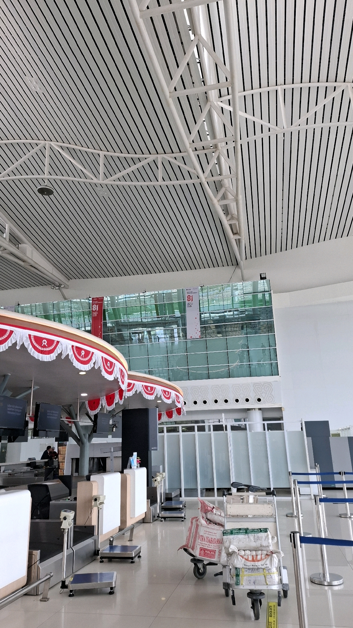 This image depicts the interior of Sultan Aji Muhammad Sulaiman Airport in Balikpapan, Indonesia. The scene is characterized by a modern architectural design with a distinctive vaulted ceiling composed of white metal slats and exposed white structural beams. Large, floor-to-ceiling windows dominate the background, reflecting the interior and the exterior environment.
In the foreground, a check-in counter area is visible. Several digital scales are positioned on the floor and counters, suggesting a baggage handling or weighing station. A luggage trolley loaded with several sacks, some bearing text in what appears to be Indonesian, is situated on the tiled floor. Decorative red and white bunting adorns the top of the check-in counters, adding a festive touch. Further into the background, frosted glass partitions create distinct zones, and blue stanchions with blue belts delineate queuing areas.
The lighting is bright, indicative of an indoor public space. A few individuals are faintly visible in the background, including one person near the check-in counters, suggesting ongoing airport operations. The overall environment is clean and orderly, consistent with a functioning airport terminal.