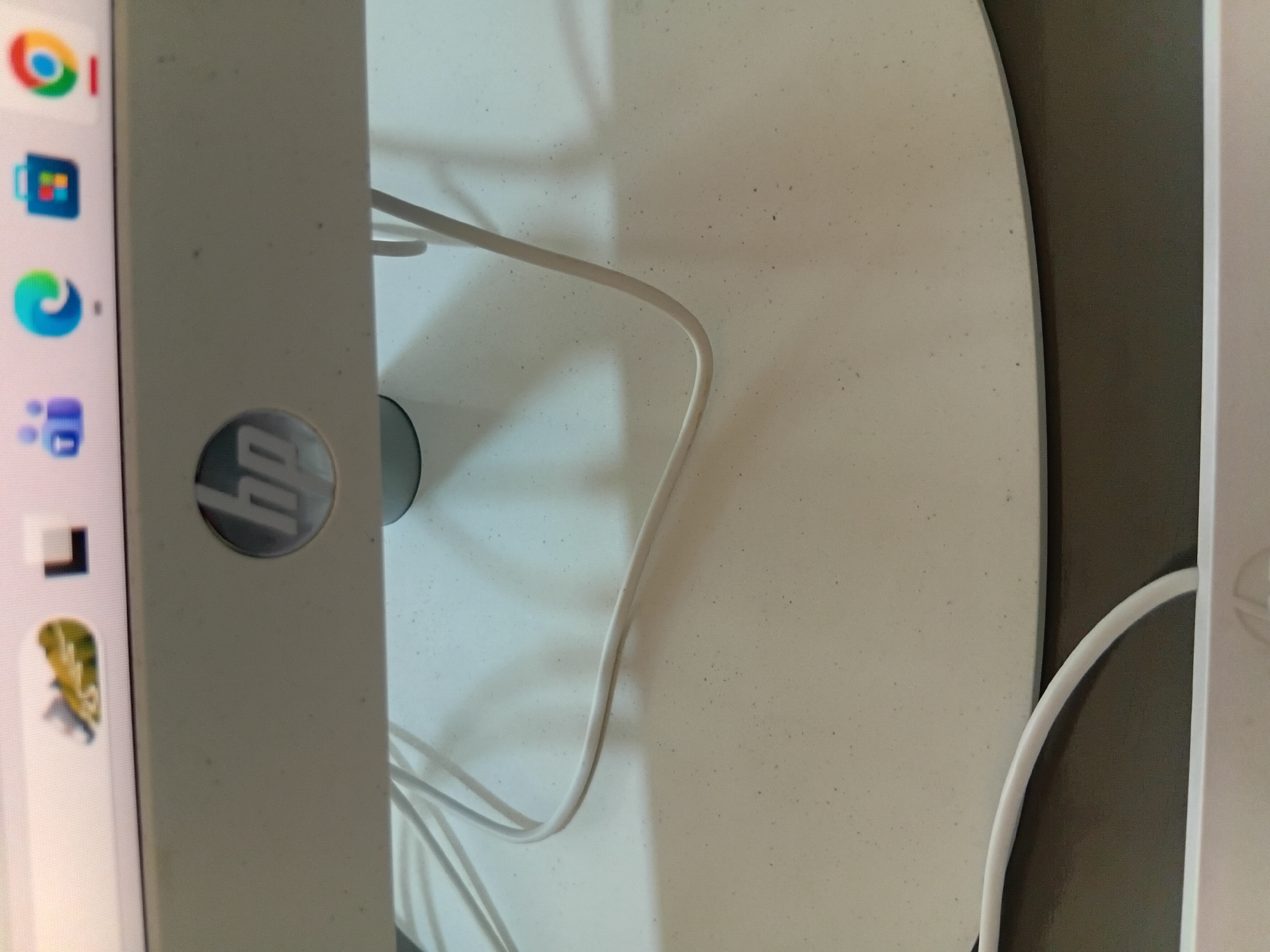 The image shows a close-up of a computer monitor's stand and base, which is white and speckled with dark dust particles. The HP logo is visible on the monitor's bezel. White cables are draped across the stand. The background is blurred, showing a wall and a portion of the computer screen displaying various icons and a web browser with a frog image. The scene appears to be indoors. There is no indication of the city of Mogadishu, Somalia in the image.