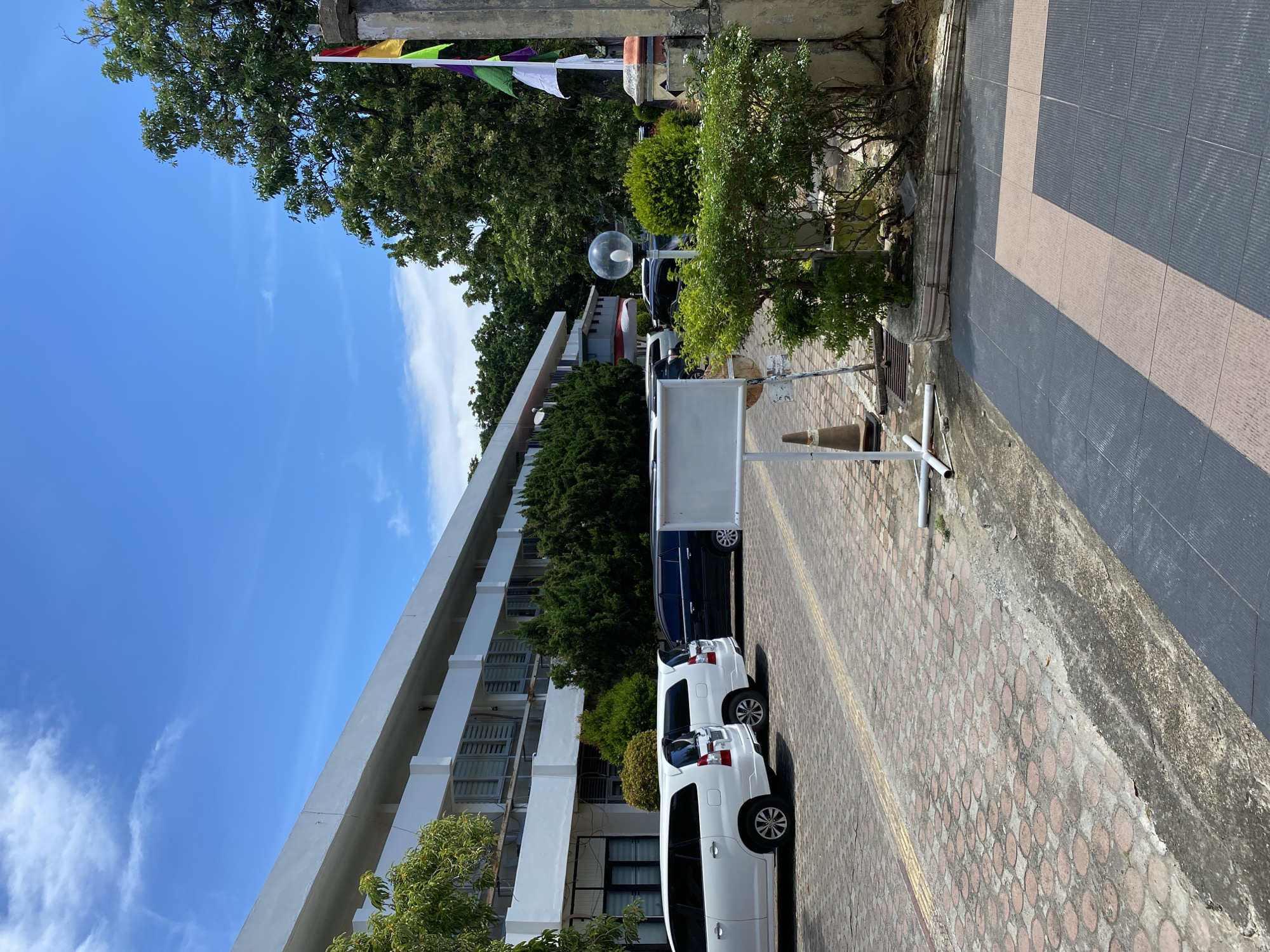 This is a photograph of the exterior of a building, likely an office or institutional building, located in Banda Aceh, Indonesia. The building itself is modern, with a long, white facade and rows of windows. In front of the building is a parking lot paved with red interlocking bricks, where several vehicles are parked, including white SUVs and a black minivan. A white portable sign on a stand is positioned in the foreground, accompanied by an orange traffic cone. Beyond the parking lot, there are various types of trees and dense green bushes, indicating a tropical or subtropical environment. A flagpole with several colorful flags is visible to the right of the frame. The sky is blue with scattered white clouds, suggesting a sunny day. The overall scene conveys a sense of order and functionality, typical of a public or commercial space.