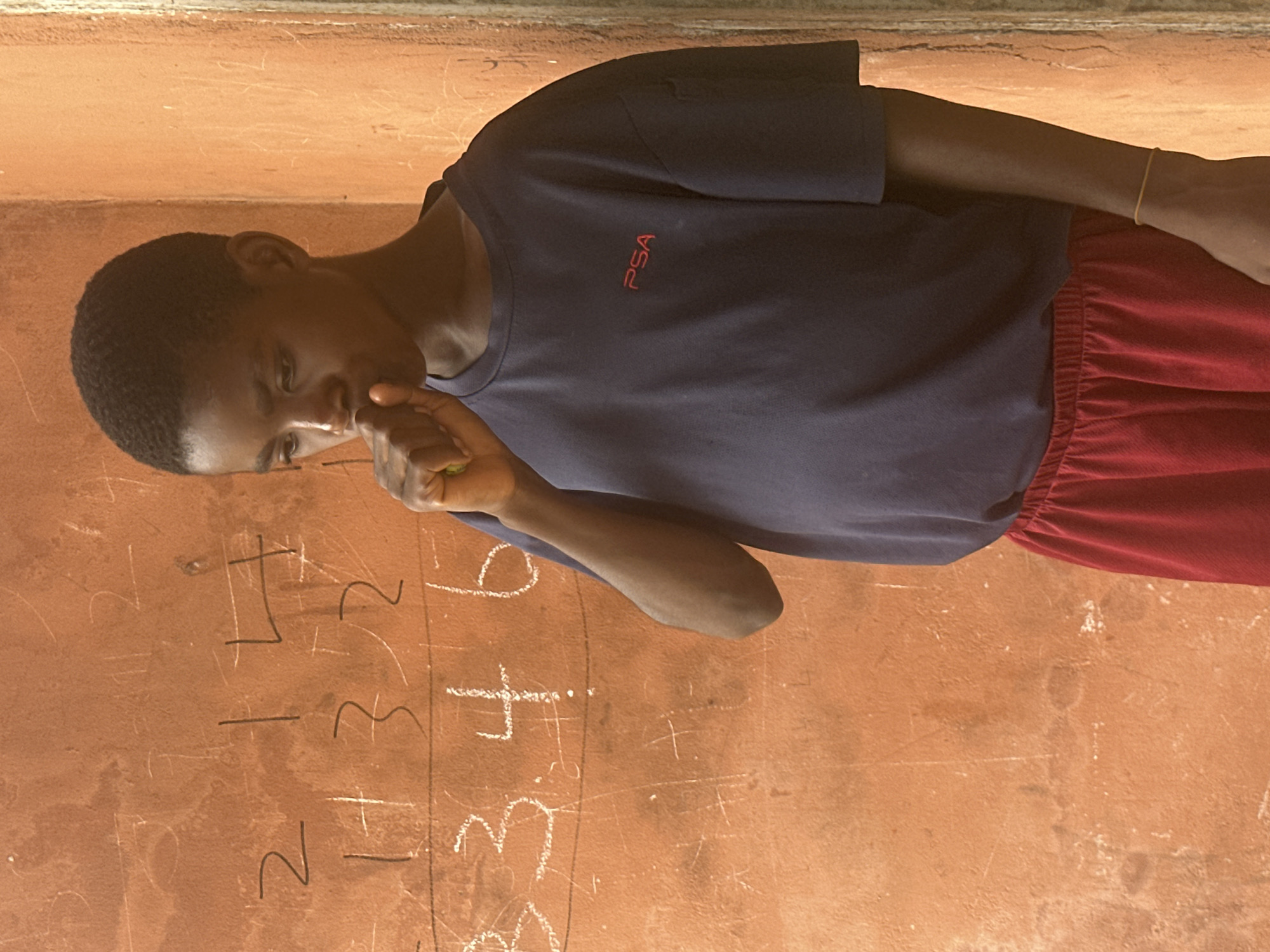 In Badagry, Nigeria, a young person stands before a chalk-covered wall with numbers and equations written on it. The person, wearing a dark blue t-shirt with a red "PSA" logo and red shorts, has their hand raised to their mouth, appearing to be thinking or perhaps coughing. The wall, an integral part of the educational environment, showcases a learning activity. The lighting suggests an outdoor or well-lit indoor setting, casting shadows that add depth to the scene. The overall impression is one of a moment captured during a lesson or study session, possibly in a school or community learning center.
