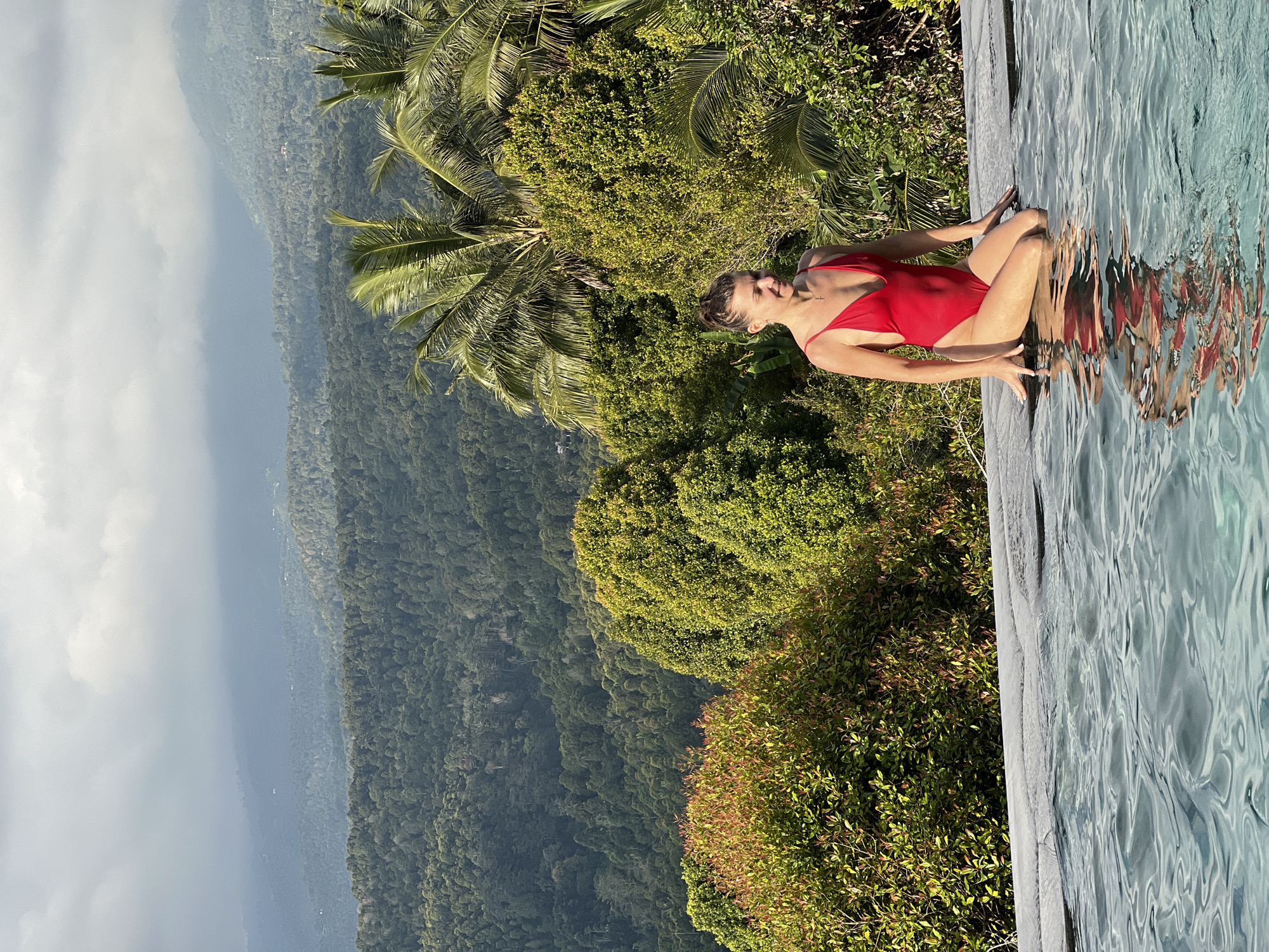A woman in a red one-piece swimsuit is kneeling on the edge of an infinity pool, her legs submerged in the water. She is looking down and smiling. The pool overlooks a lush, green landscape of rolling hills covered in dense forest and dotted with palm trees. The sky above is overcast with dramatic clouds, and the overall impression is one of serene natural beauty. The location is in Busungbiu, Indonesia.