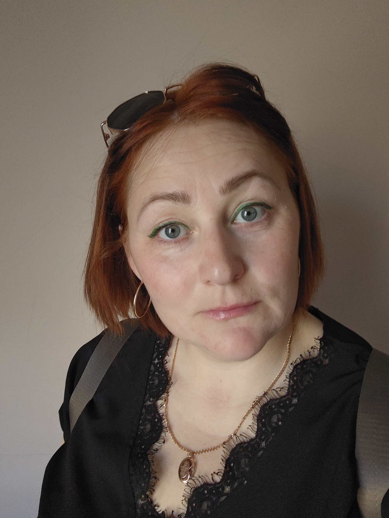A close-up, indoor photograph features a red-haired woman with green eyeliner and blue eyes, wearing a black lace-trimmed blouse and a gold necklace with a round pendant. Sunglasses are perched atop her head. She wears gold hoop earrings. A grey strap crosses her torso, suggesting a bag or purse. The background is a plain, light beige wall. The location is unknown.