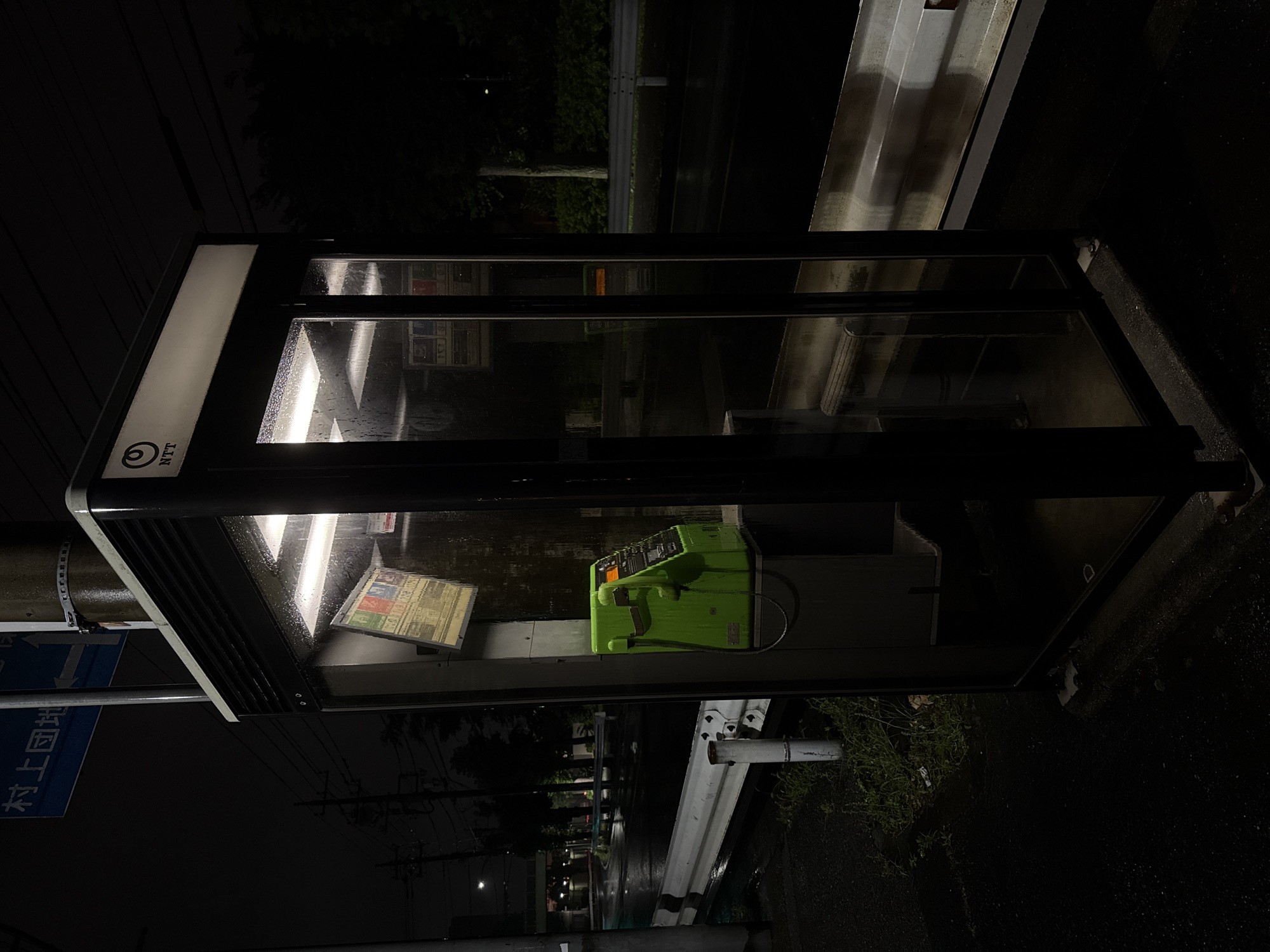This image shows a brightly lit, glass-enclosed NTT payphone booth at night in Yachiyo, Japan. The booth's interior is illuminated by fluorescent lights, casting a warm glow on the rain-streaked glass. Inside, a lime green payphone is visible, along with some papers or flyers on a shelf. The booth is situated on a wet street, suggesting it has recently rained or is currently raining. The surrounding environment is dark, with trees and streetlights visible in the background, creating a somewhat solitary and reflective atmosphere. A street sign with Japanese characters is also partially visible, confirming the location. The overall mood is quiet and perhaps a little melancholic, given the nighttime setting and wet weather.