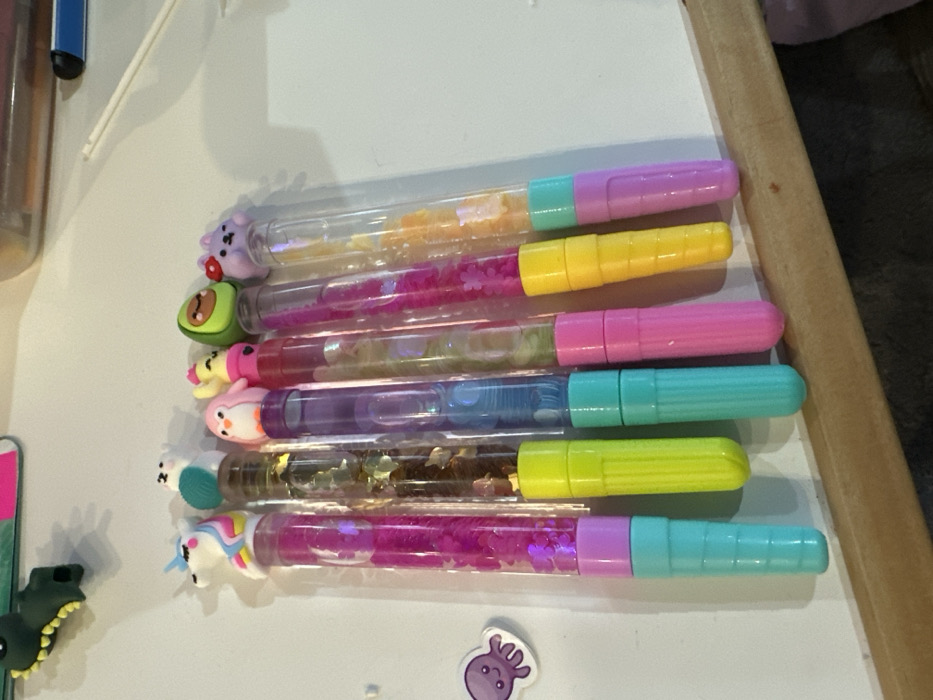 Six decorative pens are positioned diagonally from upper left to lower right on a white surface, their decorative toppers facing upwards. From left to right, the pens are characterized by their clear barrels containing liquid and glitter, topped with various figures:
1. A white unicorn topper above a barrel of pink liquid with star-shaped glitter, connected to a light purple and teal segmented base.
2. A white avian figure (possibly a bird or polar bear) wearing a teal scarf, above a barrel of gold glitter, with a neon green base.
3. A pink penguin topper above a barrel of blue liquid, with a teal segmented base.
4. A yellow chick-like figure with a pink accessory, above a barrel of light-colored liquid and glitter, with a pink segmented base.
5. A green avocado topper above a barrel of pink liquid with flower-shaped glitter, with a yellow segmented base.
6. A light purple bunny-like figure with a red mushroom on its head, above a barrel of orange glitter, with a light purple segmented base.
To the far left of the pens, a small purple octopus sticker or charm rests on the white surface. In the upper left background, a dark green, textured object resembling a dinosaur or crocodile figure is partially visible. A faint outline of a transparent container and a thin white stick are present in the upper right background. The lower edge of the image reveals a light brown wooden surface. The scene is located in the city of Varna, Bulgaria.