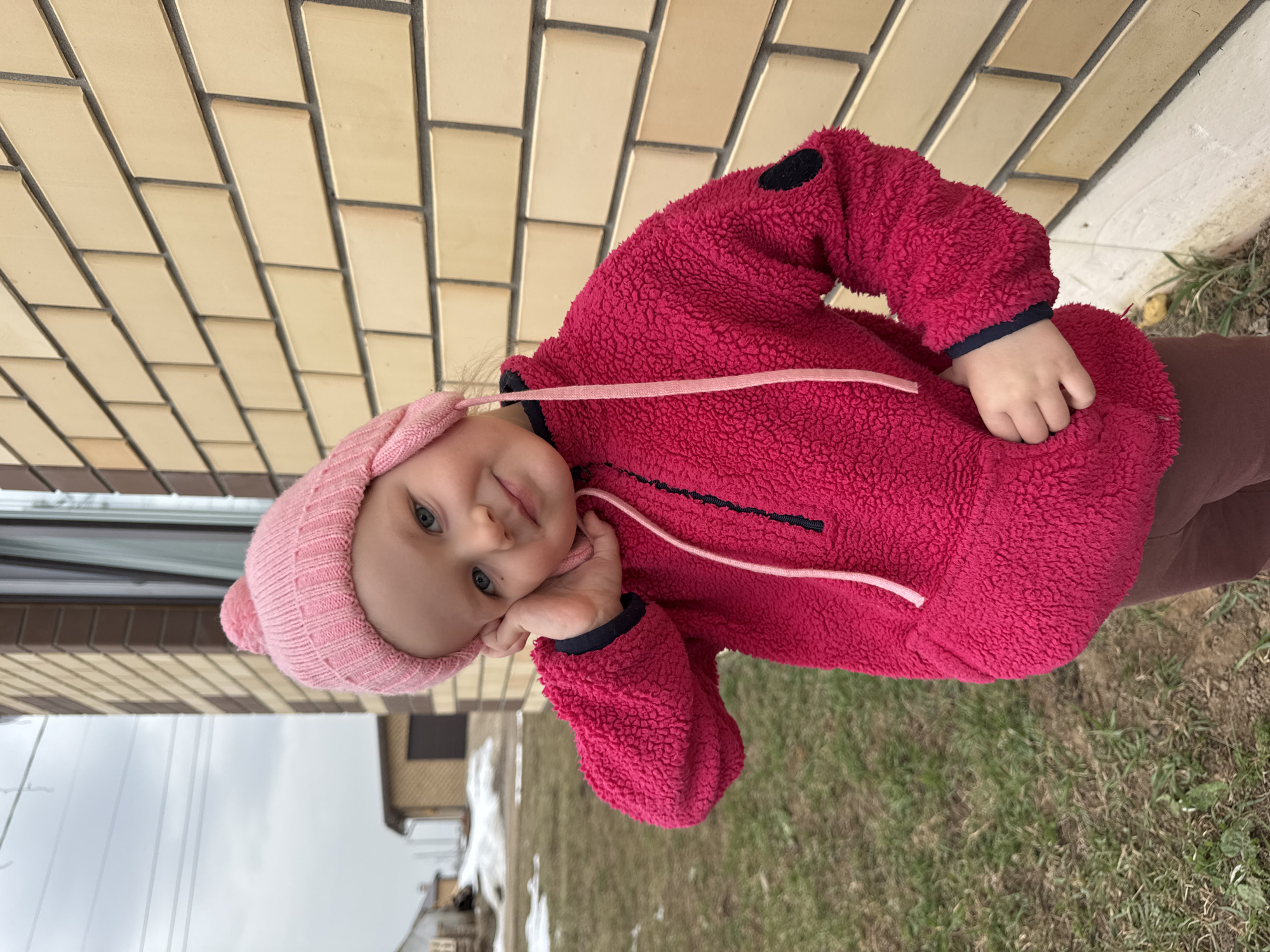 A young child, possibly a toddler, is the central subject of the image. The child is wearing a pink knit hat and a bright pink, textured fleece jacket with dark blue trim at the cuffs and a pink drawstring. The child's head is tilted slightly, and they rest one hand on their cheek. The child is standing outdoors.
The location appears to be near a building with light-colored brickwork and a window partially visible in the background. The ground shows patches of grass and possibly some remnants of snow or ice, suggesting a cool or early spring season. The scene is in Dvurechenskoe S/P in the city of Gornyi, Russia.