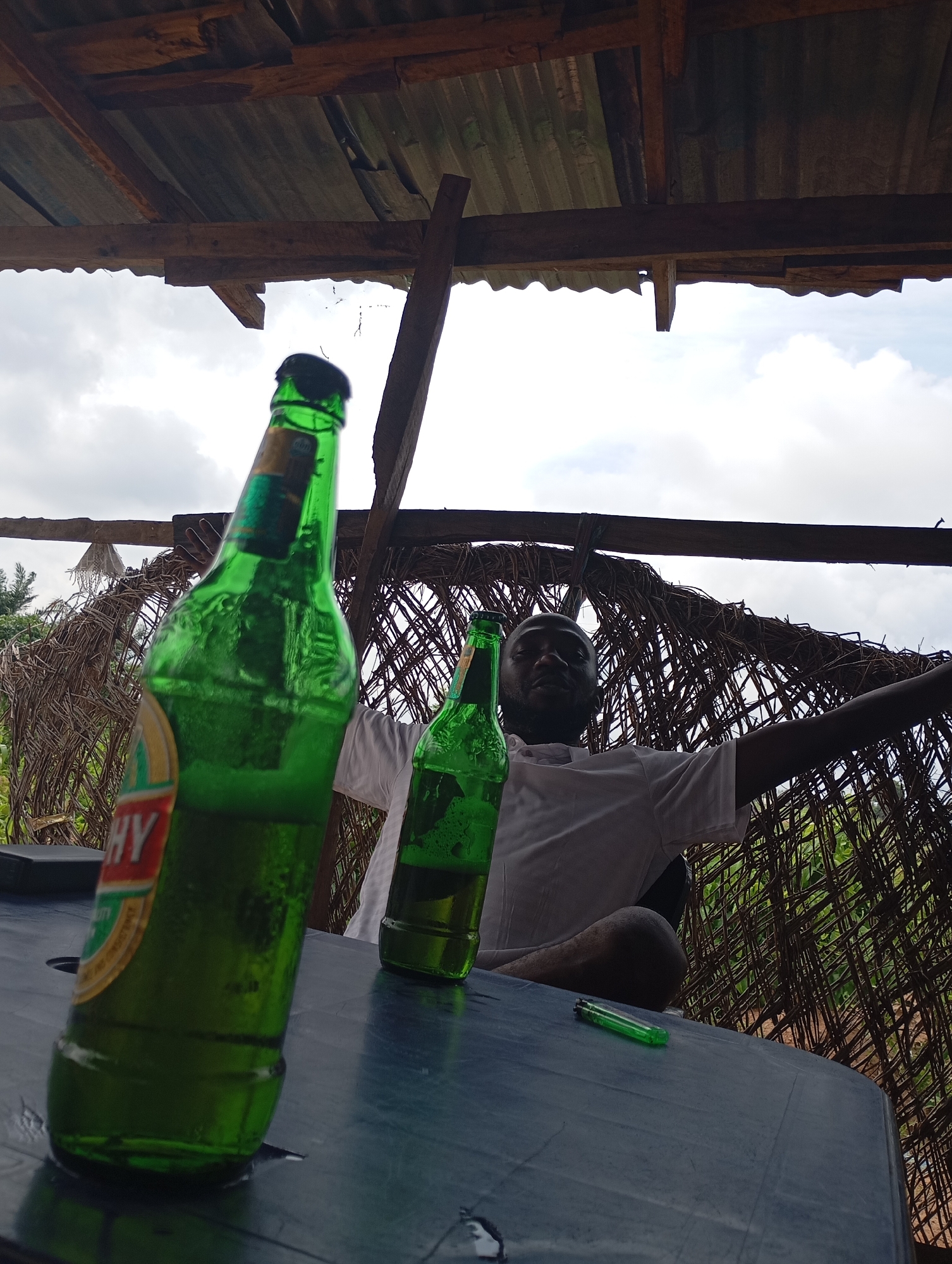 In Shendam, Nigeria, under a rustic thatched roof supported by wooden beams, a man sits at a dark table. Two green glass beer bottles, one prominently in the foreground with condensation, and another slightly further back, are placed on the table. A green lighter rests on the table between the bottles and the man. The man, wearing a white t-shirt, is partially obscured by the foreground bottle but appears to be gesturing or reaching out. The background consists of dense, woven natural materials, suggesting an open-air structure, with glimpses of greenery beyond. The sky above is overcast. The image is captured from a low angle, emphasizing the bottles and the table.