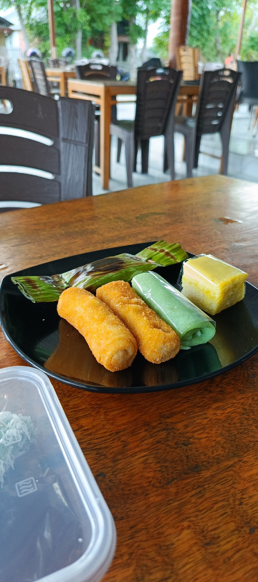 In Banda Aceh, Indonesia, a black plate rests on a wooden table, showcasing a selection of Indonesian snacks. Two golden-brown, cylindrical fritters, likely "risoles," are positioned on the left. To their right, a bright green rolled snack, possibly "dadar gulung," is visible, its creamy filling peeking out. Behind these, a portion of a snack wrapped in green pandan leaves, possibly "arem-arem," is seen. Finally, a slice of pale yellow cake, resembling "kue lapis," sits on the right edge of the plate. In the lower left corner, a clear plastic container holds a portion of a light-colored, shredded snack. The background reveals a blurred outdoor seating area with dark plastic chairs and wooden tables, suggesting a restaurant or cafe setting.