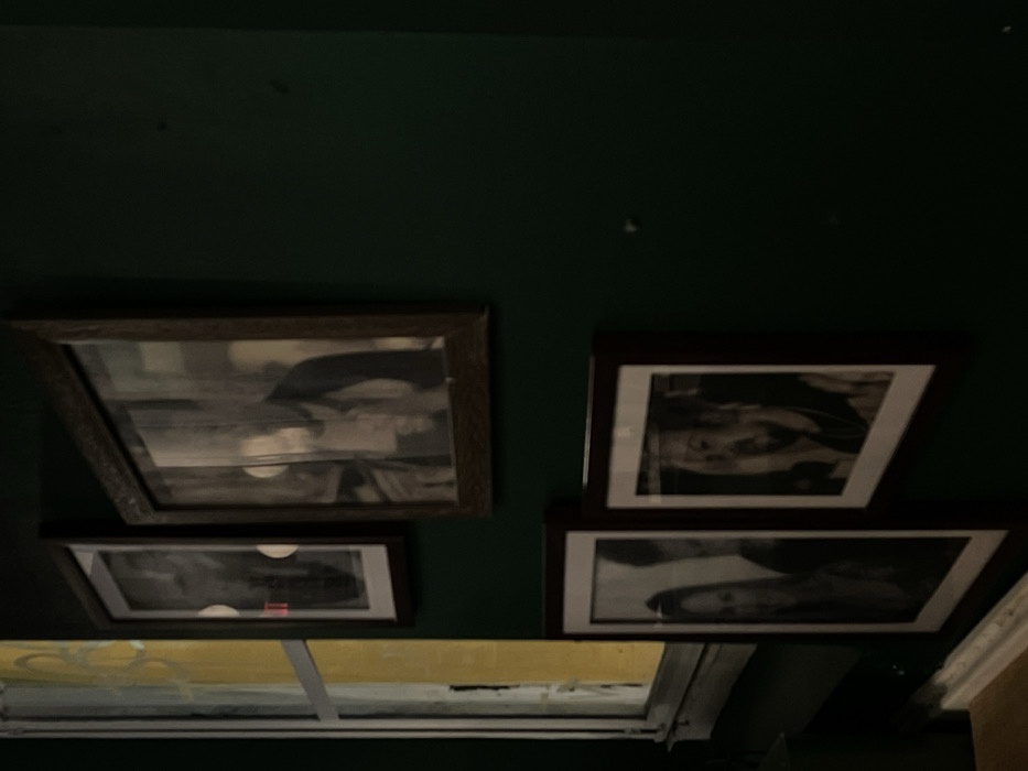 Four framed black and white photographs hang on a dark green wall. Two frames on the left have a rustic wooden finish and the right frames are a dark brown. A bright yellow and white grid ceiling is partially visible below the photos. The lighting is dim, and the image is slightly blurred.