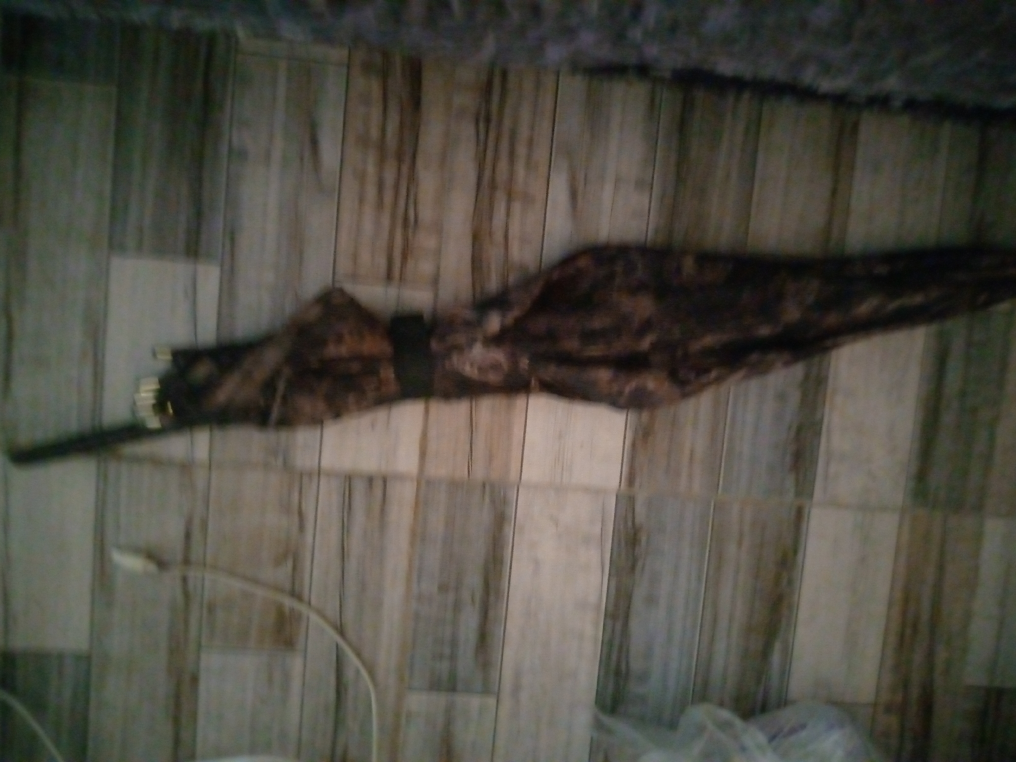 The image depicts a close-up, horizontal view of a closed umbrella lying on a wooden floor. The umbrella has a dark, patterned fabric. A thin, white cable is visible to the left of the umbrella, curving upwards and disappearing out of frame. The floor appears to be made of light-colored wooden planks with darker vertical grain lines. The overall lighting is dim, and the image is slightly out of focus. The location of the scene within Jalingo, Nigeria, cannot be determined from the provided image.
