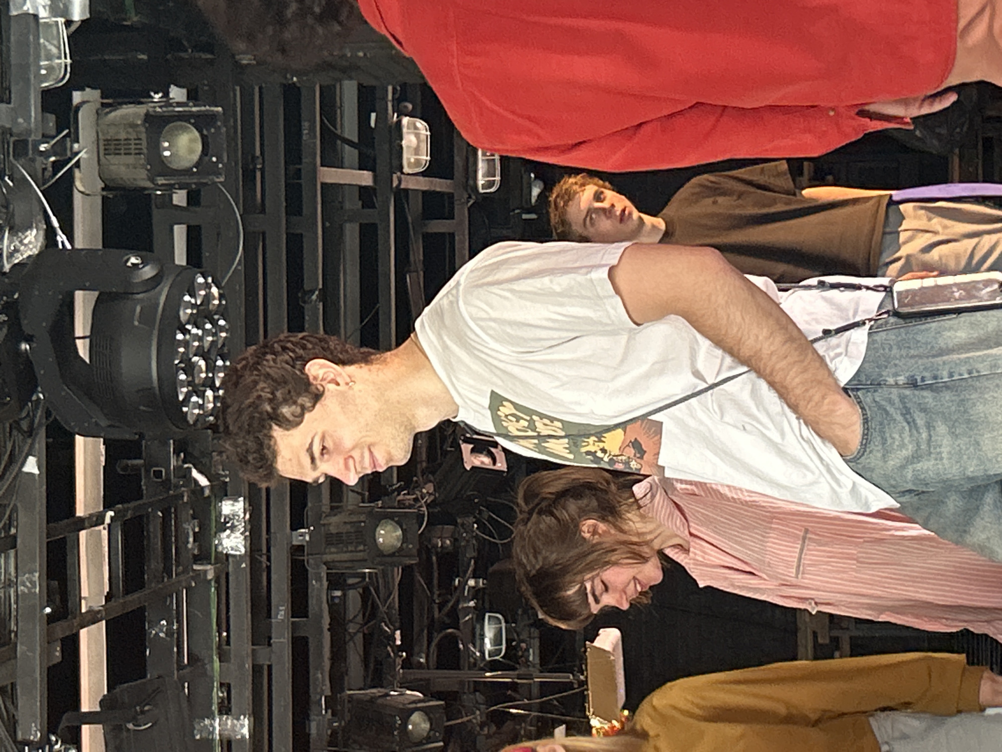 The image shows an indoor scene with four people and lighting equipment. The people are dressed in casual attire, including a white t-shirt, red sweater, brown t-shirt, pink striped shirt, and brown jacket. The lighting equipment is mounted on a metal structure. The exact location is unknown.