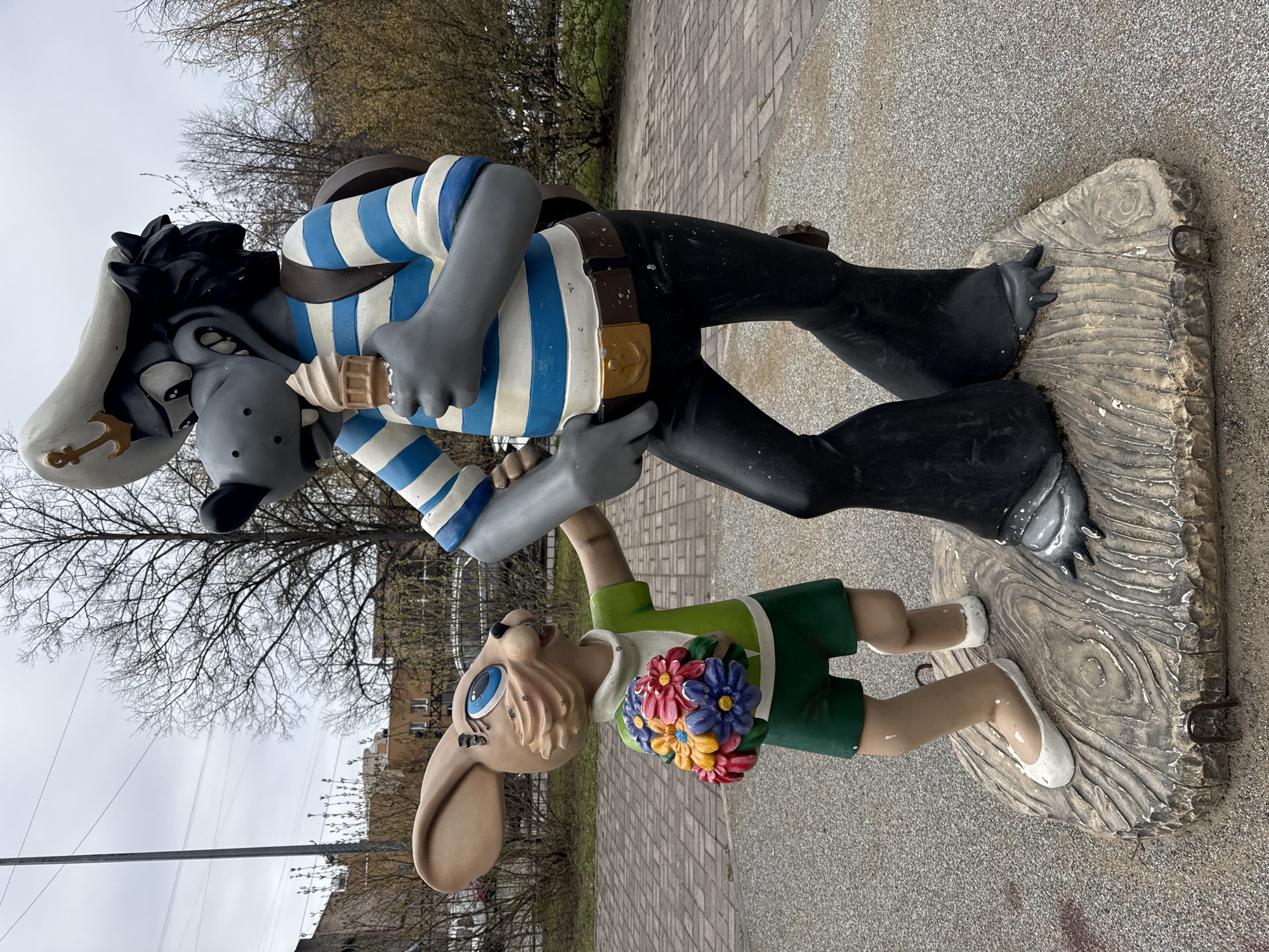 The image displays two colorful statues depicting the iconic Soviet-era cartoon characters, Wolf (Volkov) and Hare (Zayats), from the animated series "Nu, pogodi!". The scene is set outdoors on a paved surface, possibly a sidewalk or public plaza, in an urban or suburban environment, consistent with the given location of Saint Petersburg, Russia.
The larger statue represents the Wolf, dressed in a blue and white striped sailor-style shirt, dark pants, a brown belt with a golden buckle, and a light-colored hat featuring a golden anchor emblem. The Wolf holds an ice cream cone in one hand and appears to be looking down at the Hare.
The smaller statue portrays the Hare, wearing a green short-sleeved shirt, green shorts, and white shoes. It holds a cheerful bouquet of red, blue, yellow, and pink flowers and gazes upwards towards the Wolf with large, blue eyes.
Both statues are affixed to a sculpted base that mimics a textured wooden plank or raft, complete with metal rings or hooks on the sides. The background reveals bare trees, suggesting a cooler season like late winter or early spring, beneath a light gray, overcast sky. A distant building with windows and overhead power lines are also visible. The statues capture a characteristic, playful interaction between the popular cartoon adversaries. There is no visible text in the image.
