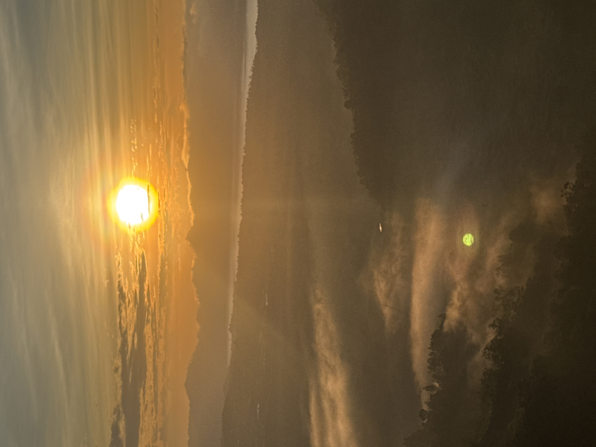This vertical, high-angle, full shot captures a breathtaking sunrise over Kintamani, Bali, Indonesia. The sun, a brilliant orb of light, sits high in the sky, casting golden rays that pierce through a thin layer of clouds. Below, a sea of mist blankets the lush green valleys and rolling hills, creating an ethereal landscape. In the foreground, trees are silhouetted against the mist, their dark forms adding depth and contrast to the scene. The reflection of the sun is visible in a small body of water nestled within the mist. The overall atmosphere is serene and majestic, conveying the beauty of a new day dawning in this picturesque location.