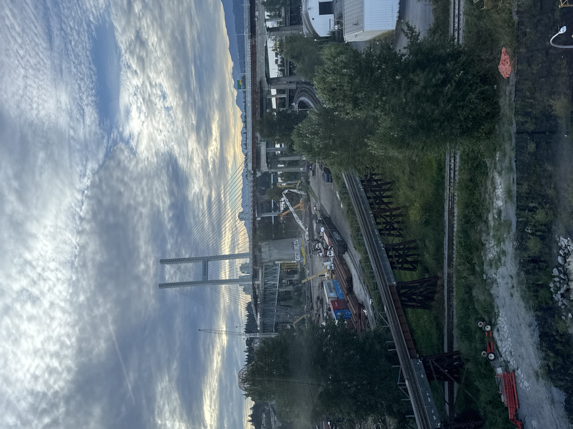 The image shows a construction site near a large suspension bridge in Surrey, Canada. The bridge, under construction, features multiple lanes and is supported by tall concrete pillars and cables. In the foreground, there are train tracks, some elevated on wooden trestles. Several shipping containers, labeled with "MAERSK" and "HANJIN," are stacked near the tracks, alongside rusted train cars and construction equipment, including cranes. The surrounding environment is a mix of industrial structures, trees, and a distant mountain range under a cloudy sky. The overall scene depicts a busy area with ongoing infrastructure development.