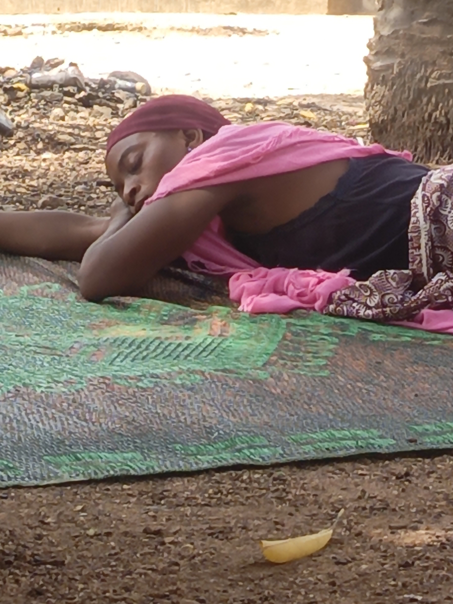 A woman is lying on a mat on the ground in what appears to be an outdoor setting. She has dark skin, is wearing a maroon head covering and a pink scarf, and is dressed in a black top. A patterned brown and white fabric is draped partially over her. She seems to be sleeping, with her eyes closed and her head resting on her arms. The mat she is lying on has a green and gray pattern. The ground is covered with dirt and small debris, with a single yellow leaf visible near the bottom of the frame. In the background, there is a pile of trash and a large tree trunk. The location is Akwanga, Nigeria.
