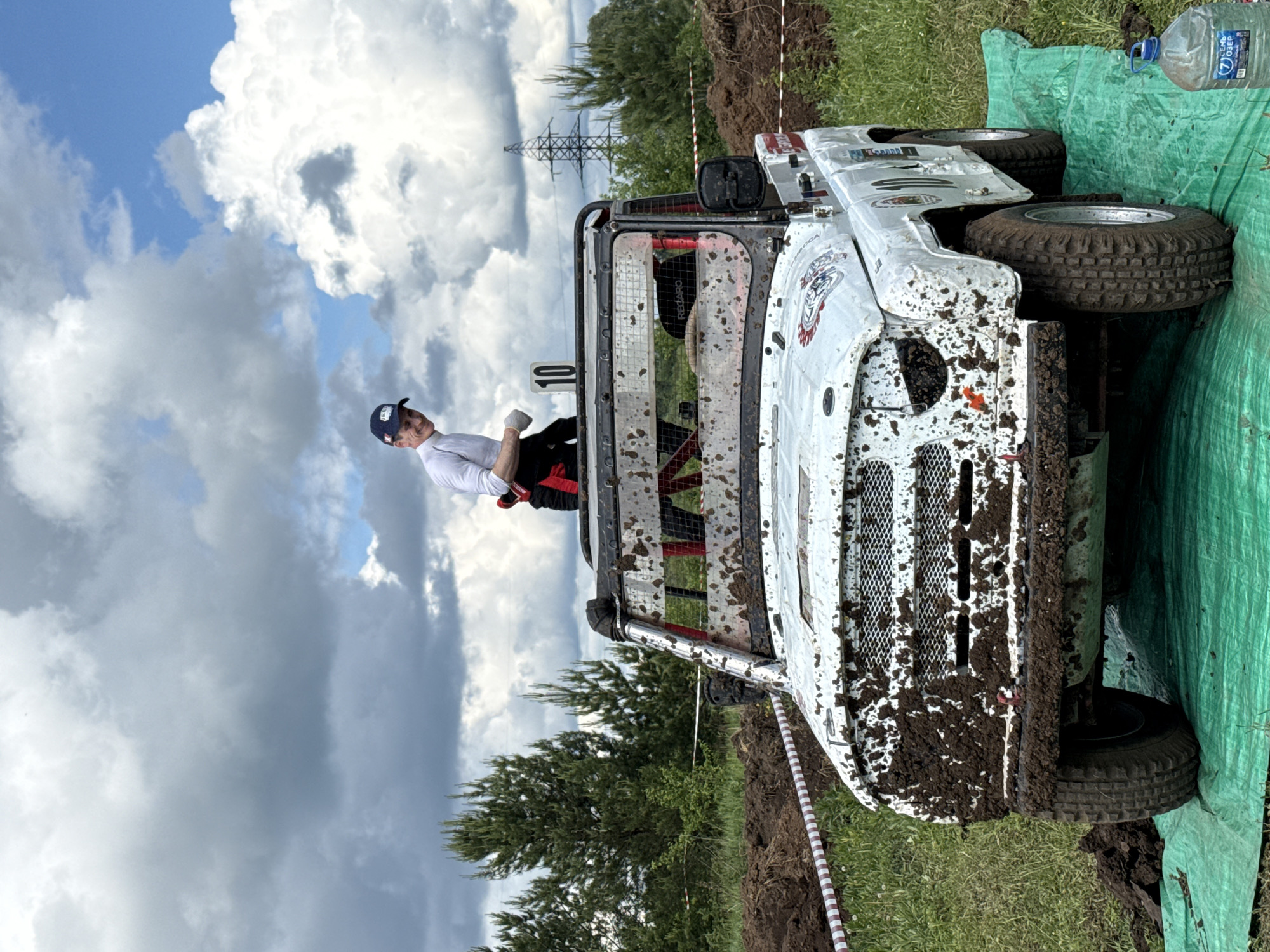 The image depicts an off-road vehicle event in Votkinsk, Russia. A white, mud-splattered buggy is partially overturned on a grassy area, partially covered with a green tarp. A man wearing a white shirt, dark trousers, a red stripe on his pants, and a blue cap is visible in the vehicle's cabin. Power lines are visible in the background. The sky is overcast with clouds. A water bottle is visible on the tarp near the vehicle. A dirt embankment is visible behind the vehicle, along with a red-and-white striped barrier.