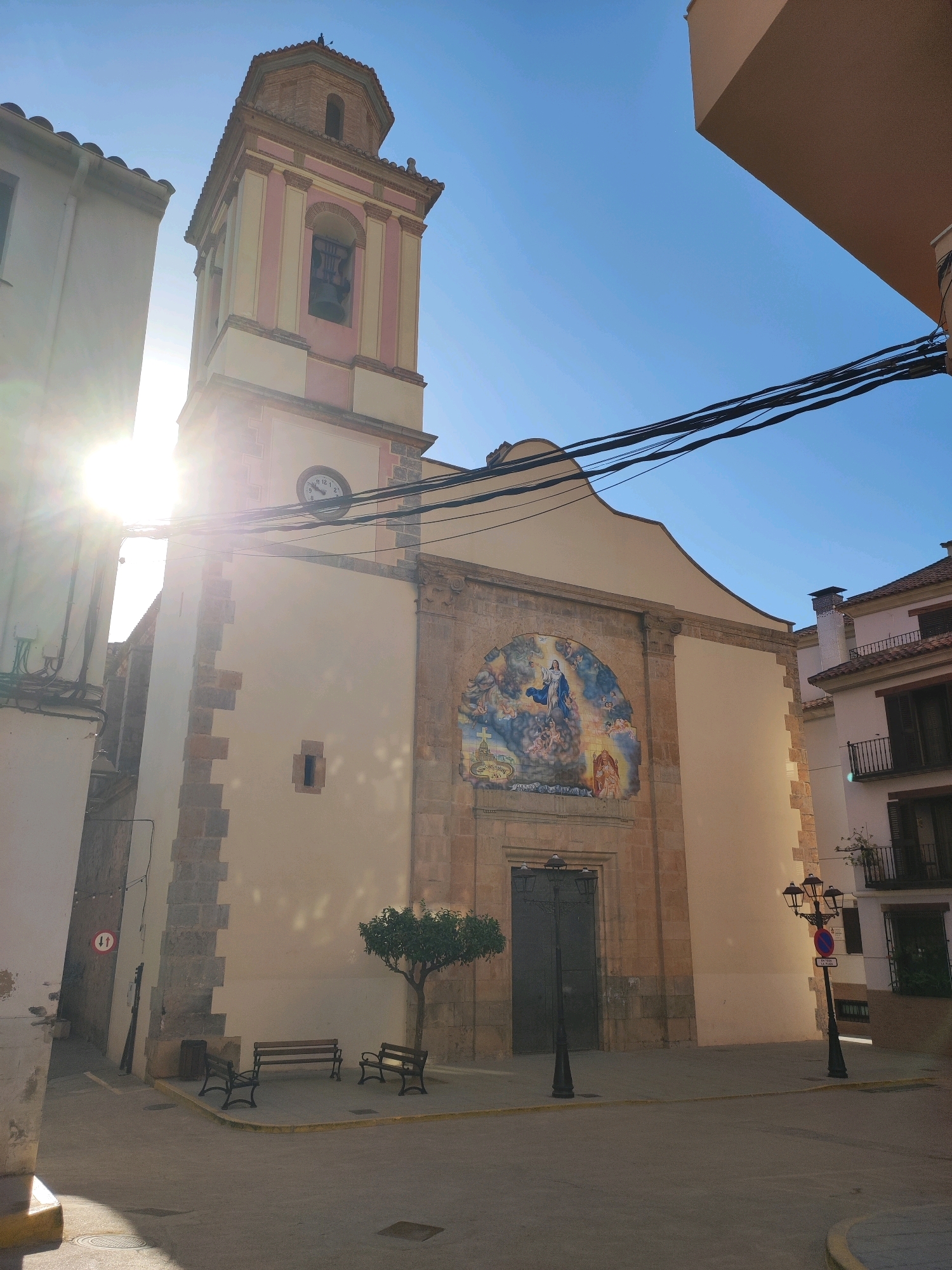 This image depicts the facade of a church in the town of Suera, Spain, under a clear blue sky. The church features a tall bell tower with a pink and yellow striped design and a clock face. Above the entrance to the church is a large, arched mural depicting a religious scene, likely the Assumption of Mary, with figures surrounded by clouds and cherubs. The church's walls are a light yellow, with stone accents around the edges and entrance. In front of the church, there is a paved public square with a small tree, two park benches, and a trash receptacle. A street lamp stands to the right of the church entrance. The scene is bathed in sunlight, with lens flare obscuring part of the bell tower. To the left of the church, a building casts a shadow onto the square. The overall environment suggests a peaceful, sunny day in a Spanish town.