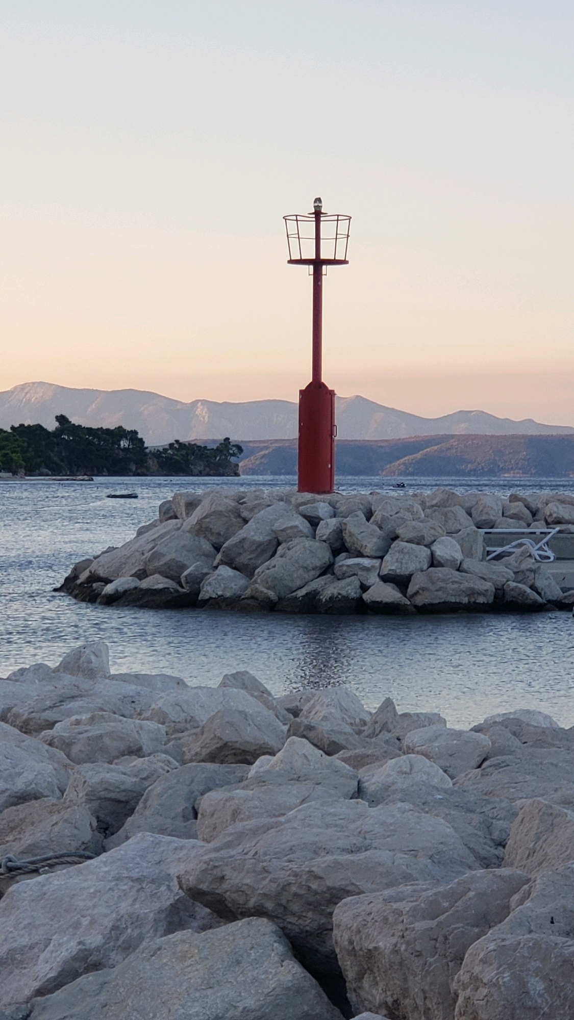 The image shows a red navigation light on a pier made of large, light-colored rocks. The pier extends into calm, blue water. In the background, there is a coastline with green trees and rolling hills or mountains under a pale, hazy sky. The scene appears to be set during either sunrise or sunset, indicated by the soft, warm light. The location is identified as Općina Tučepi, Croatia. There are no people visible in the image. The overall atmosphere is peaceful and serene.