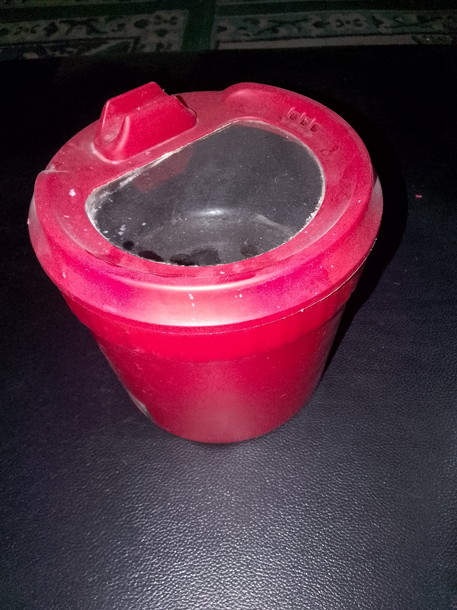 A red plastic container with a transparent lid sits on a textured dark surface. The lid has a small raised handle and some faint markings. The container appears to be a storage item. The background is out of focus, showing a dark surface with a patterned object above. The image is taken from a slightly elevated angle, looking down at the container.