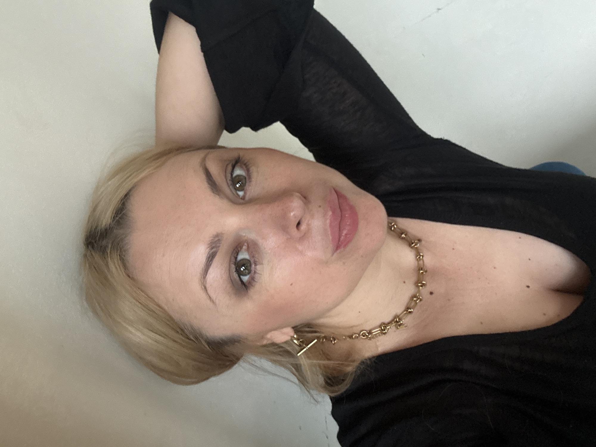 A blonde woman with light skin, brown eyes, and fair makeup poses for a selfie against a light-colored wall. She is wearing a black top with a low neckline and a gold necklace. Her left arm is bent, with her hand resting on the back of her head, and her right arm is visible extending down her side. She is also wearing gold earrings. The image shows a close-up of her face, with her gaze directed towards the camera. There is no information to determine the location in the city of Sochi, Russia.