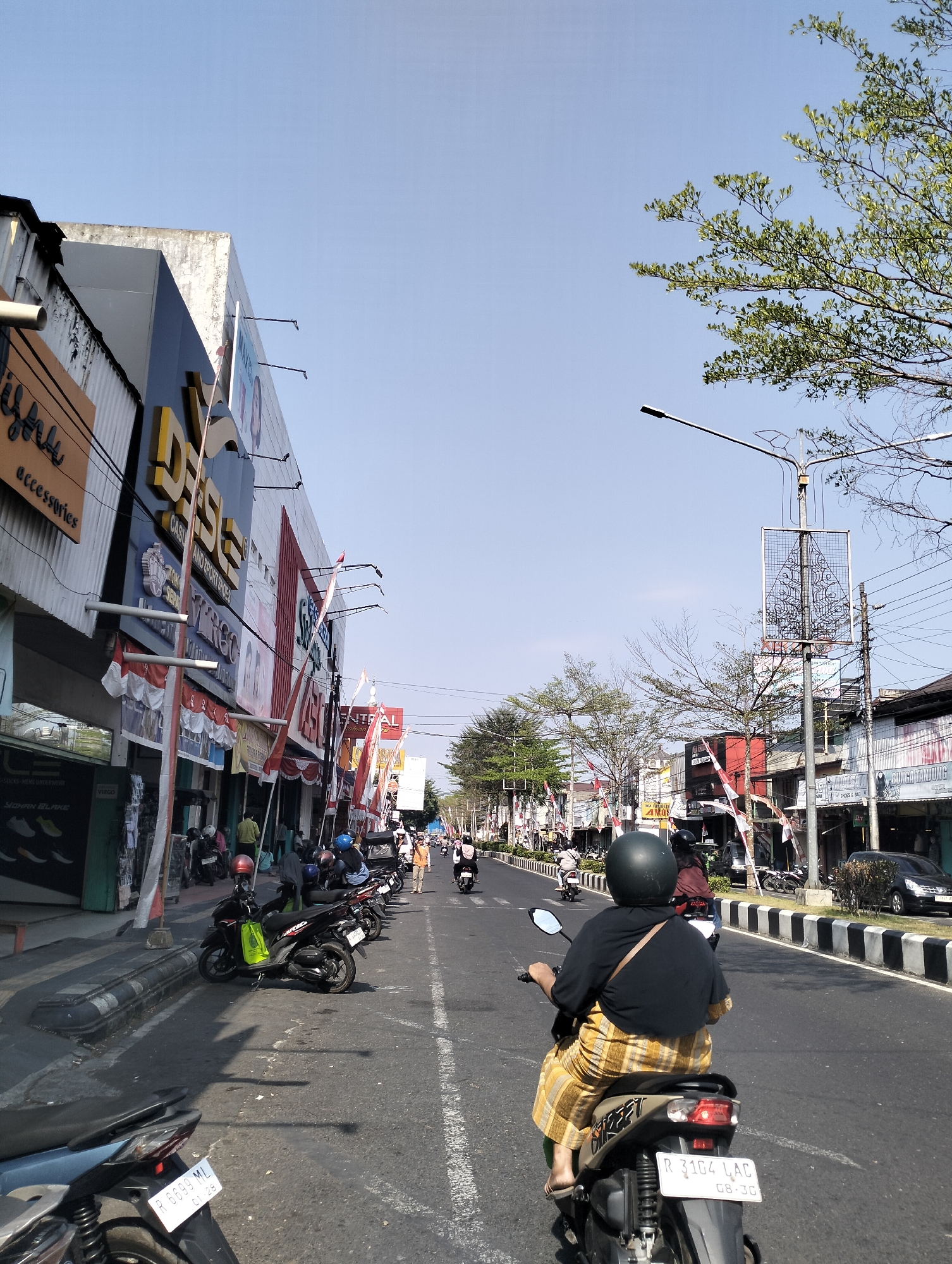 The image depicts a street scene in Purbalingga, Indonesia, characterized by a clear blue sky and bright daylight. A road runs through the center of the frame, with traffic consisting of several motorcycles and a few cars. The foreground features the rear of a person riding a scooter, wearing a dark shirt and a checkered yellow and brown skirt, along with a dark green helmet. The scooter has a license plate visible, displaying "R 3104 LAC 08-30". To the left of the road is a row of buildings with storefronts. One prominent building displays a large sign with yellow lettering that appears to spell "DESGO" followed by other text. Indonesian flags are visible, hanging from poles along the street. The right side of the road also has buildings and a sidewalk with trees lining it. The overall atmosphere suggests a typical day in an urban environment with moderate commercial activity.