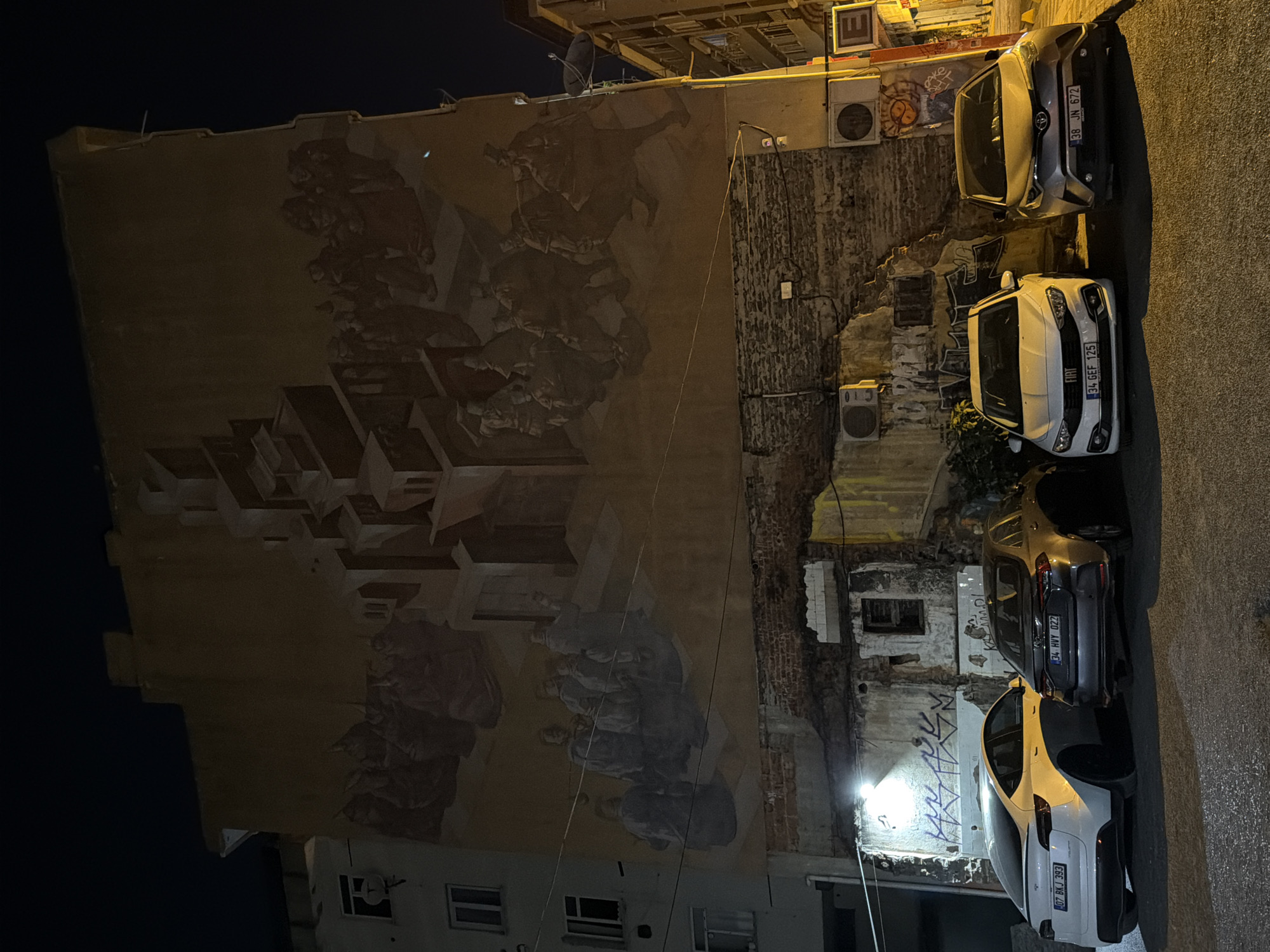 The image shows a night scene in the city of Kadıköy, Turkey, with a large mural painted on the side of a building. The mural depicts a surreal, tiered architectural structure surrounded by figures in historical attire. In the foreground, four cars are parked: a white Tesla Model Y, a silver sedan, a white Fiat sedan, and a silver hatchback. The building behind the cars appears aged and partially dilapidated, with exposed brickwork and graffiti. Lighting in the scene comes from streetlights and the headlights of the cars.