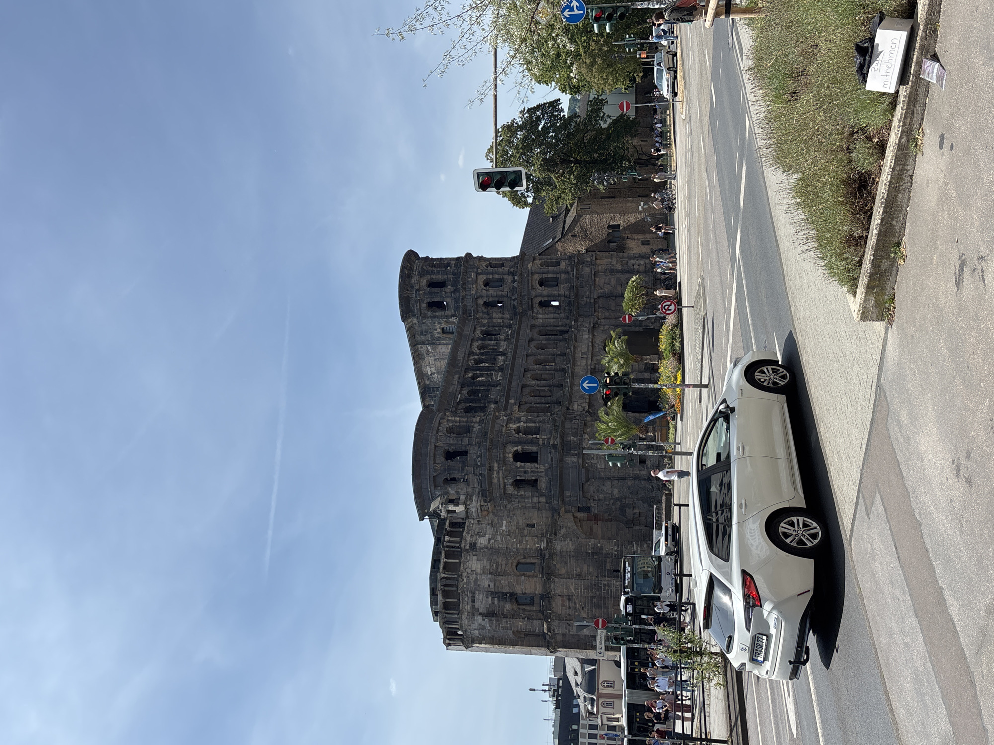 The image captures a street scene in Trier, Germany, focusing on the Porta Nigra, a remarkably preserved ancient Roman city gate. In the foreground, a white station wagon is parked on the right side of a crosswalk. To the left, a busy street features a white bus with passengers boarding or disembarking, and several pedestrians walking along the sidewalk. The Porta Nigra dominates the background, a massive stone structure with multiple levels and arched windows, displaying evident signs of age. The sky above is a clear, bright blue with a few wispy clouds. Traffic lights are visible, one showing a red signal. Lush green trees and some flowering plants add color to the urban environment. Several traffic signs, including directional arrows and prohibition signs, are strategically placed. The overall impression is of a sunny day in a historic city square, with the ancient monument serving as a prominent landmark.
