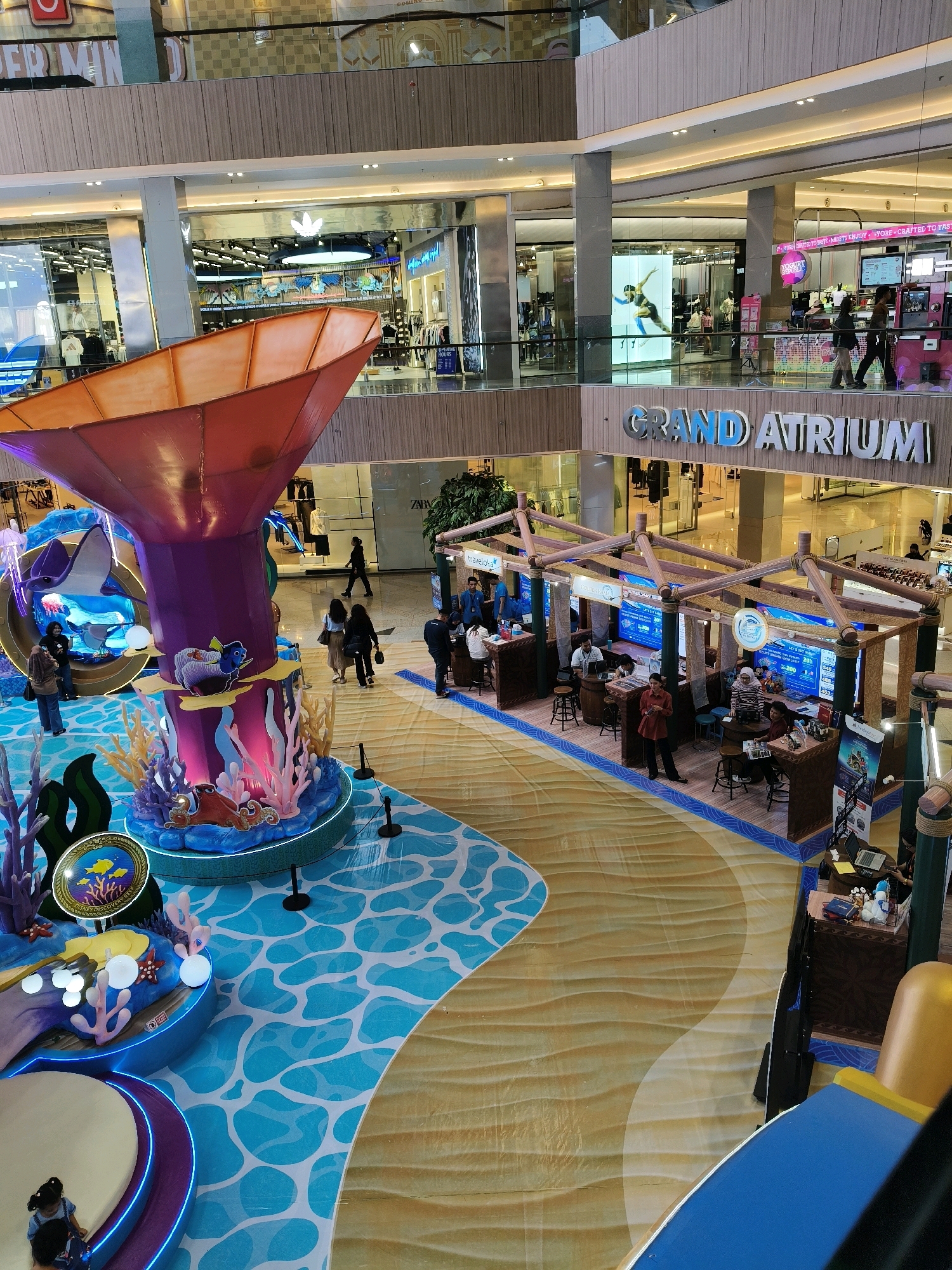 The image shows an interior view of a multi-level shopping mall, identified by the "GRAND ATRIUM" signage. The mall features several retail stores with glass storefronts, including what appears to be an Adidas store with its logo visible. In the foreground, there is a large, decorative display themed around an underwater or ocean scene, featuring colorful coral, sea creatures like an octopus and a stingray, and stylized water patterns on the floor. Several people are visible throughout the mall, some walking, some standing or interacting with stalls. One stall, positioned under a wooden canopy, seems to be an information or sales booth with screens displaying promotional content and staff interacting with customers. The lighting suggests an indoor environment, likely during the day. The architecture includes escalators, railings, and the typical elements of a modern shopping complex. The presence of "GRAND ATRIUM" signage and the style of the mall are consistent with locations in Surabaya, Indonesia.