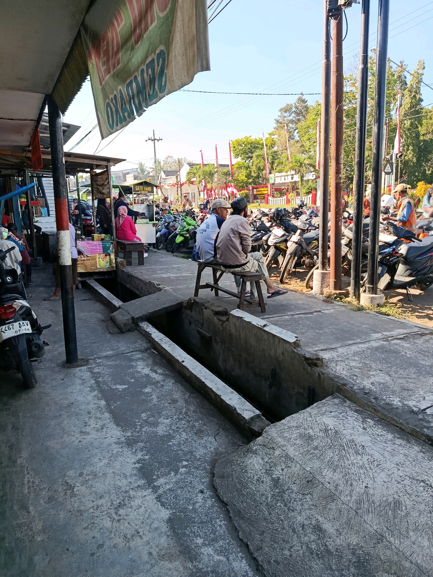 A bright, sunny day captures a street scene in Bululawang, Indonesia. In the foreground, a motorcycle is parked on a concrete sidewalk. The sidewalk features a drainage ditch, partially covered by concrete slabs. Several people are seated on benches and stools, some conversing while others appear to be waiting. Behind them, a row of motorcycles is parked. Further back, a food stall labeled "SATE TAHU" is visible, with customers and vendors around it. The background shows more parked motorcycles, a street with a few cars, trees, and buildings decorated with Indonesian flags, suggesting a national holiday or celebration. The overall impression is of a busy, typical day in a local Indonesian town.