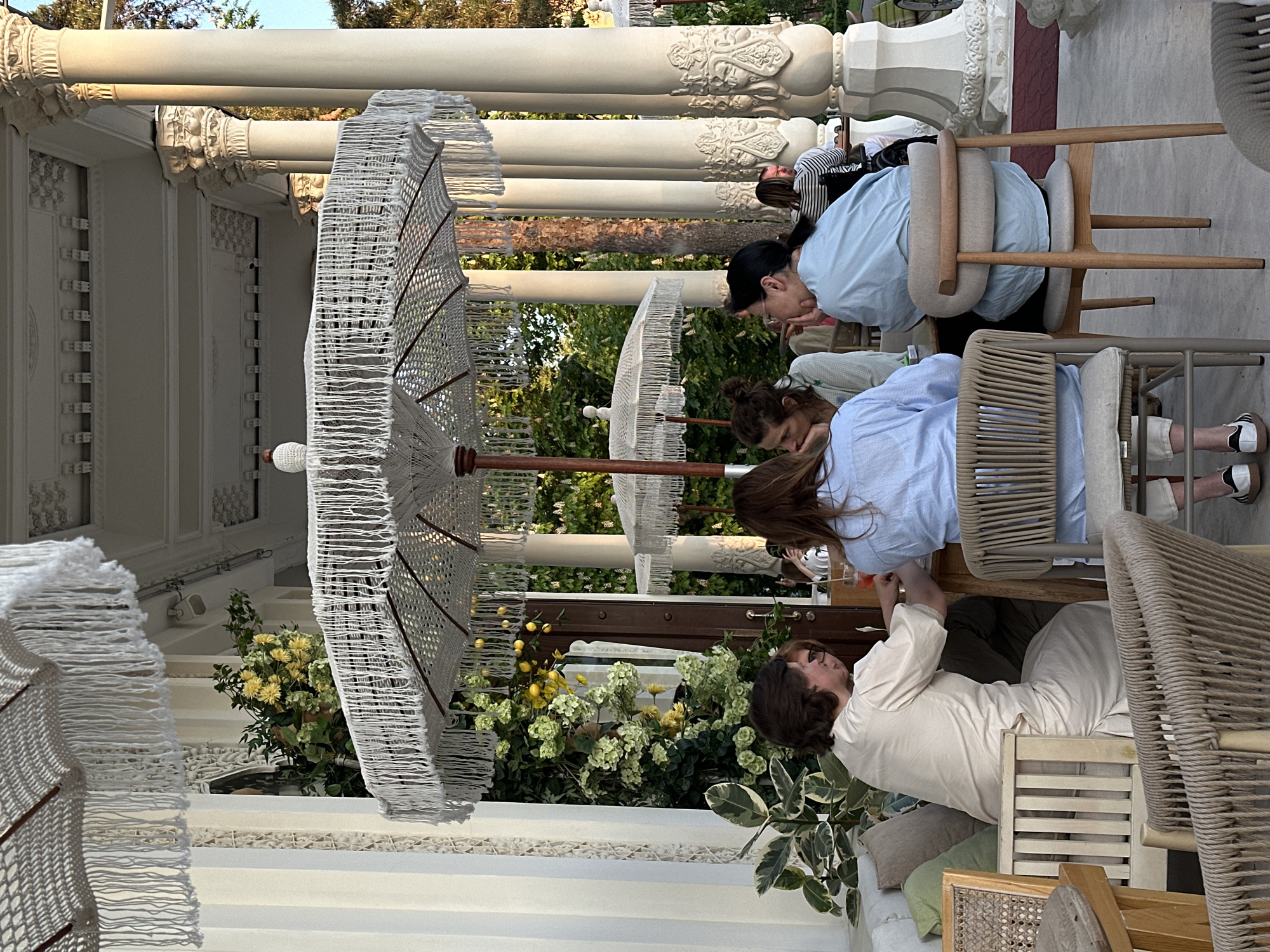 The image shows an outdoor dining area in an unidentified city. The architectural style suggests a Mediterranean influence with ornate white columns and detailing. Several white fringed parasols provide shade to the tables. Three women are seated at a table, engaged in conversation or activity. One woman is dressed in a light beige top, another in a light blue shirt, and a third in a darker blue shirt. The table is set with drinks, and various plants, including lemon trees and flowering shrubs, decorate the area. The furniture is a mix of wood and woven materials.
