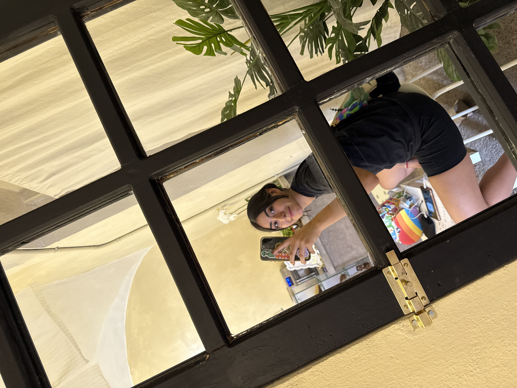 This is a mirror selfie of a young woman, captured through a dark-framed mirror or window. The image is rotated, presenting the woman and her reflection in an inverted orientation.
The main subject is a young woman, seen from the waist up, smiling pleasantly and looking directly at the camera. She has dark hair and is wearing a dark t-shirt and dark shorts. She holds a smartphone with a colorful, decorated case (featuring what appears to be a green alien and other cartoon-like elements) to take the photo.
The reflection reveals parts of an indoor setting. Behind the woman, elements of a room are visible, including a chair with white legs, a low table (possibly glass), and a rainbow-colored item, which could be a cushion or bag. A large green plant, resembling a Monstera, is also reflected in the upper panes of the mirror/window.
The mirror or window itself is characterized by a prominent black frame, divided into a grid pattern. The frame shows signs of wear and tear, with some chipped paint revealing older material underneath. On the bottom right section of the frame, a golden-colored metal bolt lock is attached, bearing the engraved text "ALEGANTULA."
Beyond the reflective surface, through the upper glass panes, a light-colored, possibly off-white, pleated fabric is visible, likely curtains or blinds, suggesting another interior space or light source. The lighting appears to be ambient indoor light. The overall scene conveys a casual, personal moment captured within a home setting in Puebla, Mexico.