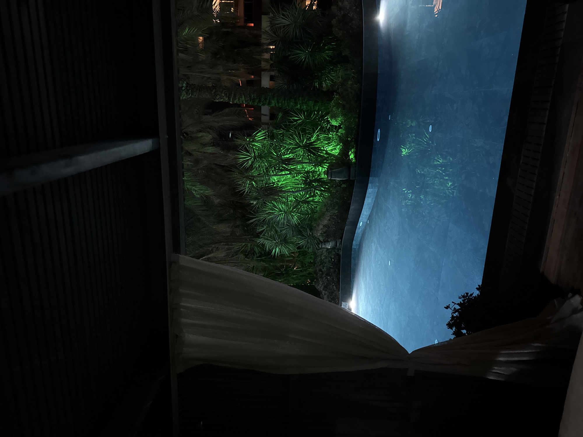 A nighttime, low-angle, wide shot shows a swimming pool in the foreground, illuminated by subtle underwater lights. Beyond the pool, lush greenery is dramatically lit with vibrant green lights, creating a striking contrast against the dark night sky. Palm trees are visible, their fronds silhouetted against the illumination. In the far background, a building with lit windows suggests an urban or resort setting. A white sheer curtain drapes into the left side of the frame, partially obscuring a dark structure, possibly a covered seating area or cabana. The scene evokes a sense of a tranquil, private oasis, likely in a warm climate.