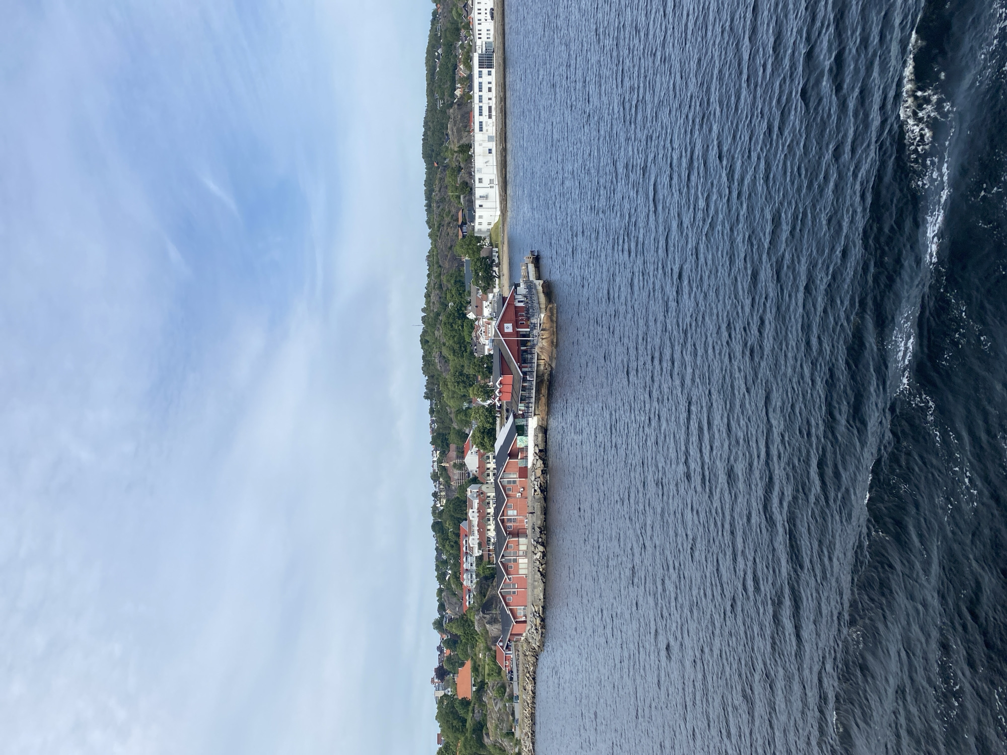 This is a rotated landscape image of a coastal town in Larvik, Norway. The scene depicts a town nestled along a rocky coastline, with buildings primarily in shades of red and white, many featuring traditional architectural elements with red roofs and white trim. The town is backed by a lush, green, tree-covered hillside.
The water body to the right of the town is a dark blue, appearing choppy with white foam and waves breaking near the shore, suggesting movement and perhaps a moderate breeze. The sky above is a pale blue with wispy, white clouds, indicating it is daytime, likely late morning or early afternoon, with fair weather conditions.
In the foreground, several houses are visible along the water's edge, some with docks. A larger white building with many windows is situated further up the hillside. The overall impression is of a picturesque, lived-in coastal community. There is no visible text in the image.