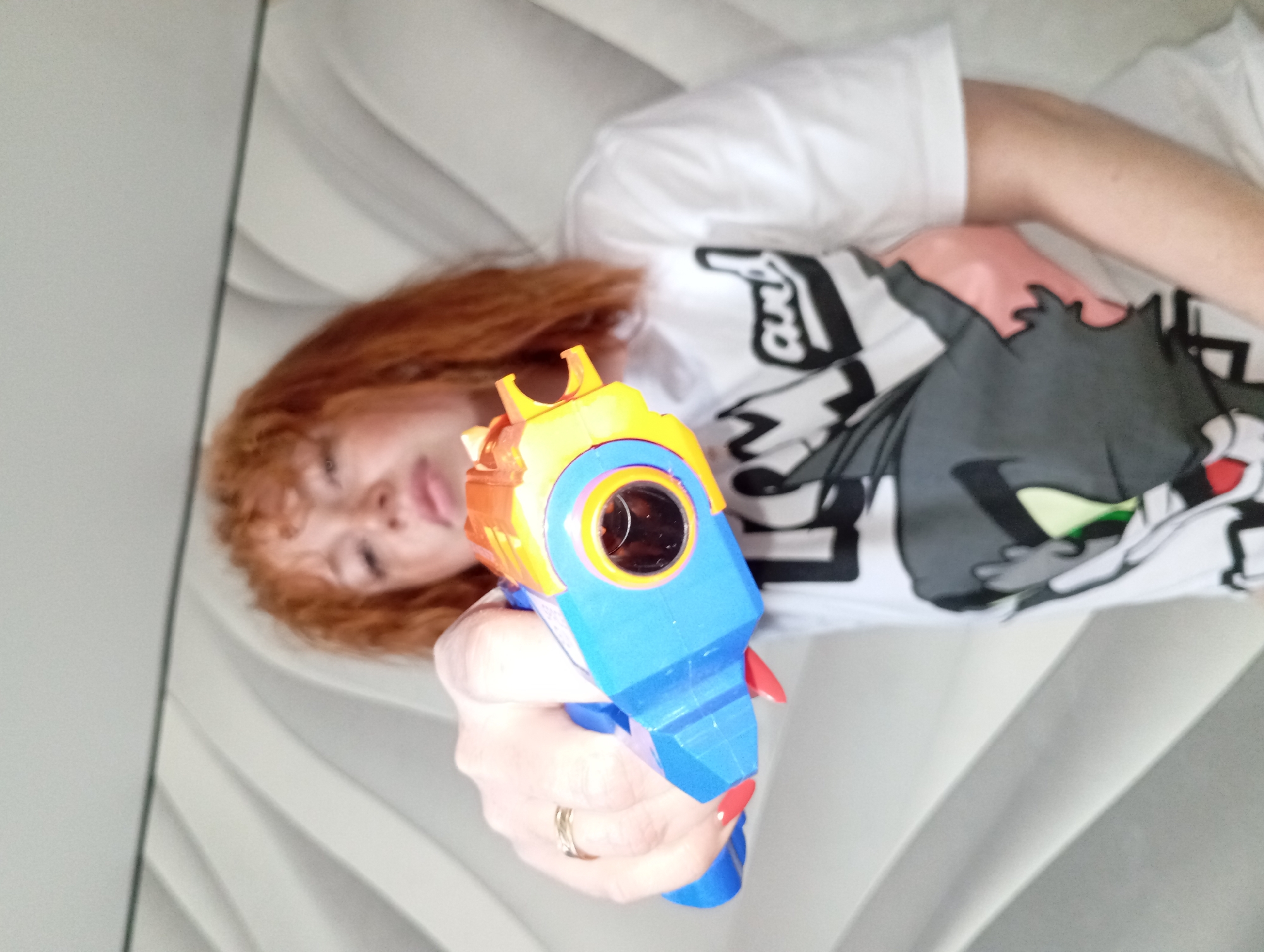 A woman with curly red hair is lying down, pointing a toy water gun directly at the viewer. She is wearing a white T-shirt with a cartoon cat graphic. Her left hand, adorned with a gold ring, is gripping the blue and orange water gun. The background features a wavy gray and white surface. The location is unknown.