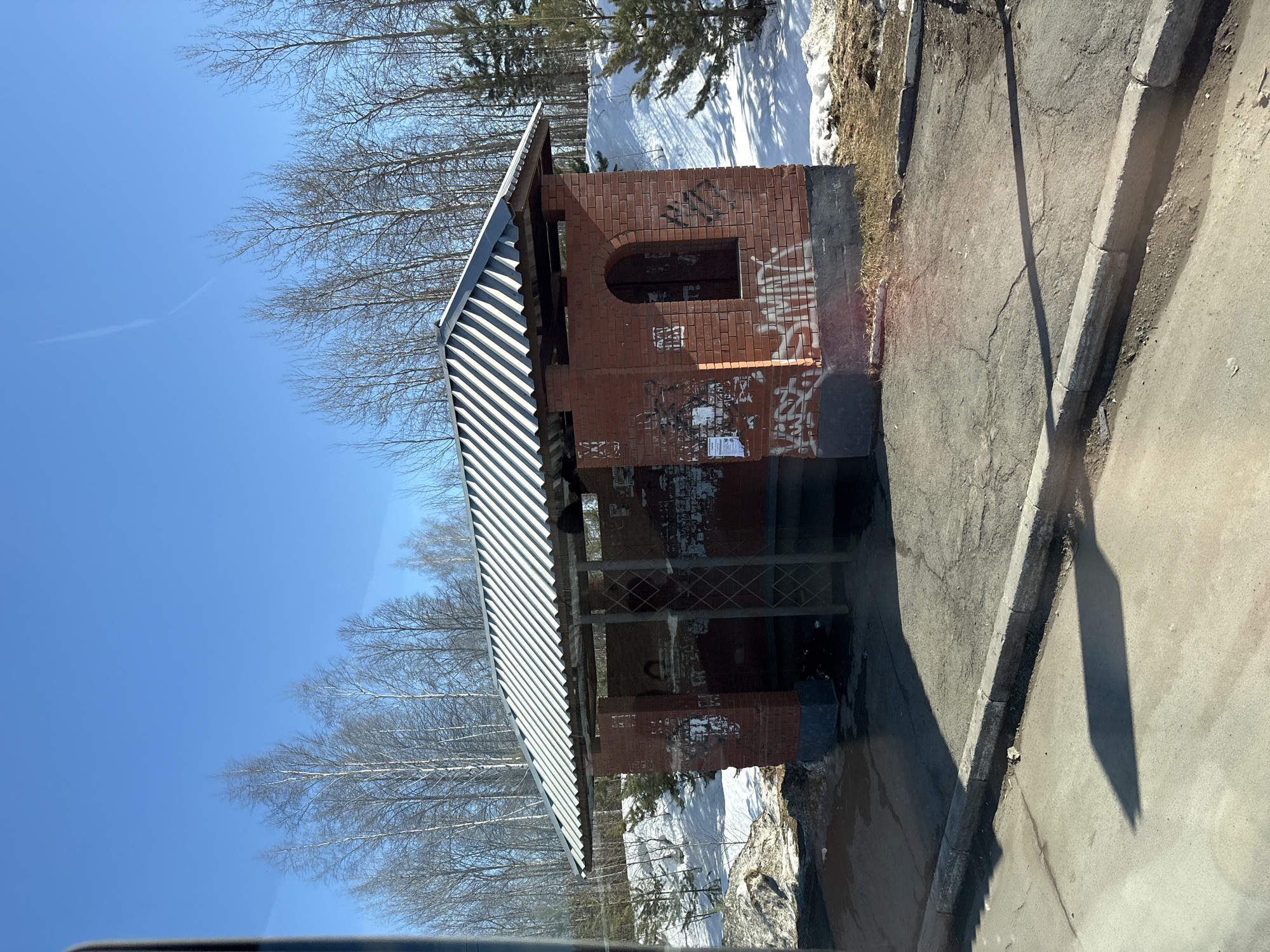 The image depicts a red brick bus stop shelter with a corrugated metal roof, situated on the side of an asphalt road in Bolgury, Russia. The structure features a dark gray or black painted base, and its red brick walls are extensively covered with white and black graffiti tags and torn paper posters. One side of the shelter shows an arched, window-like opening. Inside the shelter, a simple frame or support structure is visible, and debris is present on the ground.
Surrounding the bus stop, patches of melting snow and exposed dry earth/vegetation are visible, suggesting a late winter or early spring season. Behind the shelter, a snow-covered embankment rises, populated by numerous bare deciduous trees, possibly birches, with a few small evergreen conifers also present. The sky above is clear and bright blue, indicating sunny weather. A distinct shadow from the bus stop structure falls across the cracked asphalt surface in the foreground, where a concrete curb separates the road from the bus stop area. No individuals or active interactions are visible within the scene.