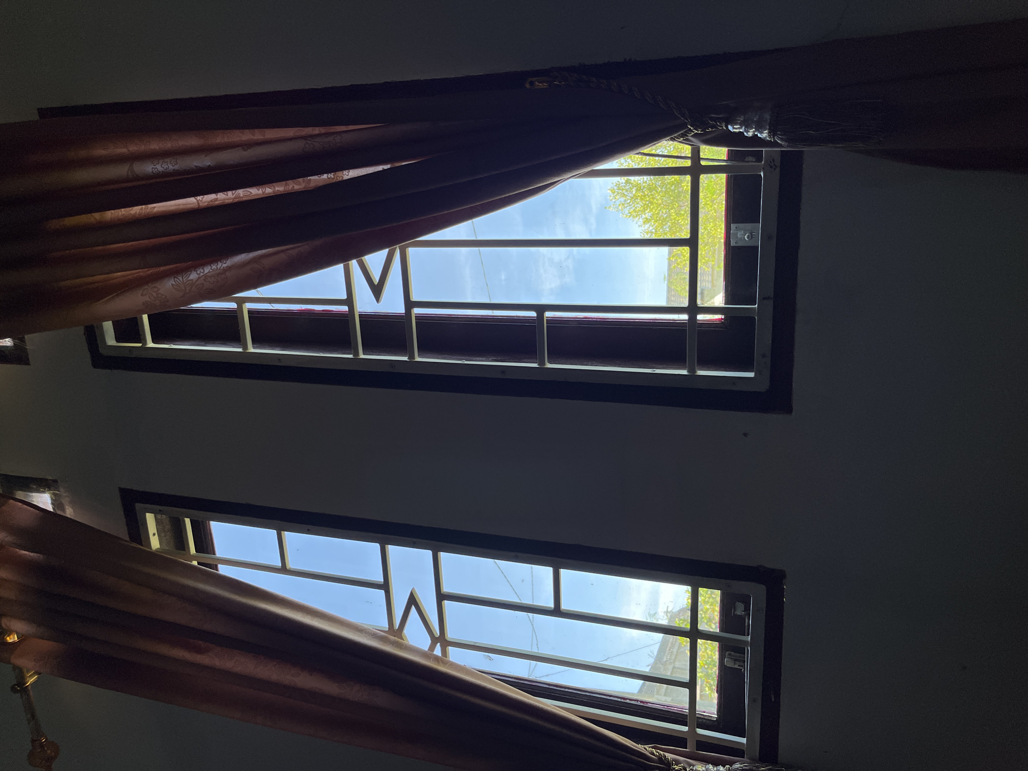 The image captures the interior of a structure in Lhokseumawe, Indonesia, featuring two tall, vertically oriented windows set into a dimly lit wall. Each window is framed in dark wood and secured with white metal grilles; the grille on the left window incorporates a prominent zigzag design in its upper section. Dark reddish-brown curtains are partially drawn, framing both windows; the curtain on the right is held by a decorative tassel tie-back.
Through the windows, a bright blue sky with scattered white clouds is visible, indicative of daytime. Lush green foliage, consistent with trees, is present at the base of the exterior view through both windows. A utility line is faintly discernible across the upper portion of the view through the left window, alongside distant, indistinct structural elements. The interior walls appear dark due to the low light conditions, creating a strong contrast with the brightly illuminated outdoor scene. No people or active interactions are depicted.