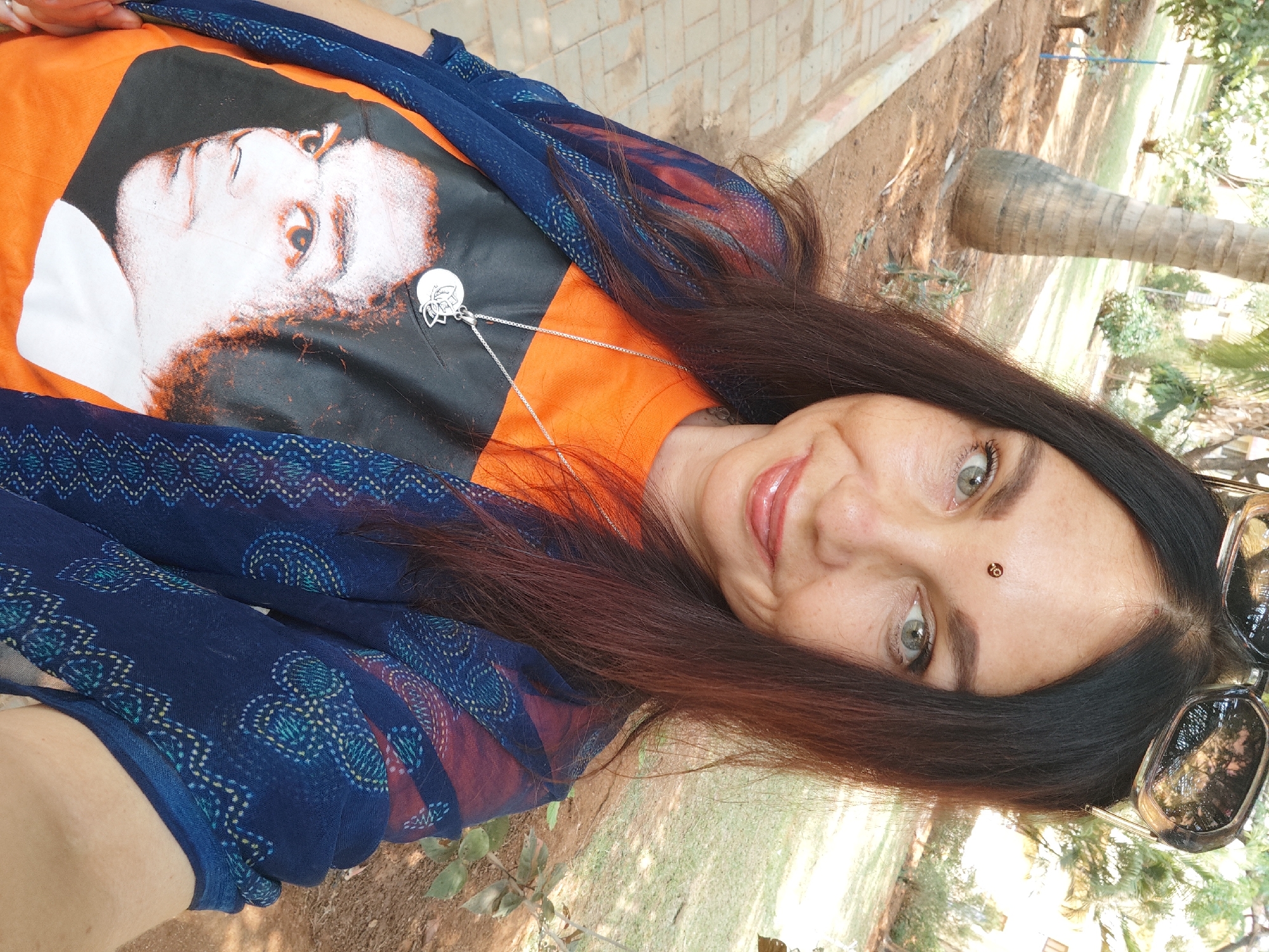 The image is a selfie of a woman with long, dark brown hair, light green eyes, and a small brown bindi on her forehead. She is wearing a bright orange t-shirt with a black and white graphic print of a face on it, along with a silver necklace featuring a pendant. She is draped in a blue patterned scarf. Sunglasses are perched on top of her head.
The background shows an outdoor environment with trees and vegetation. The ground is covered in dirt and grass. There is a section of a tree trunk visible. The city and specific location are not discernible from the image.