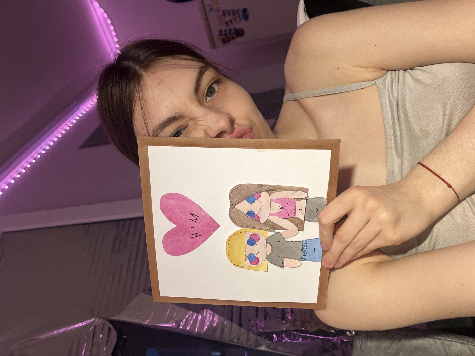 A young woman is holding up a framed drawing, while posing for a picture. The drawing features two stylized figures, one with brown hair and a pink top, and the other with blonde hair and a grey shirt. Above the figures is a pink heart with the letters "H+M". The woman is wearing a light grey tank top and has a red string bracelet on her wrist. A string of purple LED lights is visible in the background. The exact location in Saint Petersburg, Russia, is not discernible from the image.
