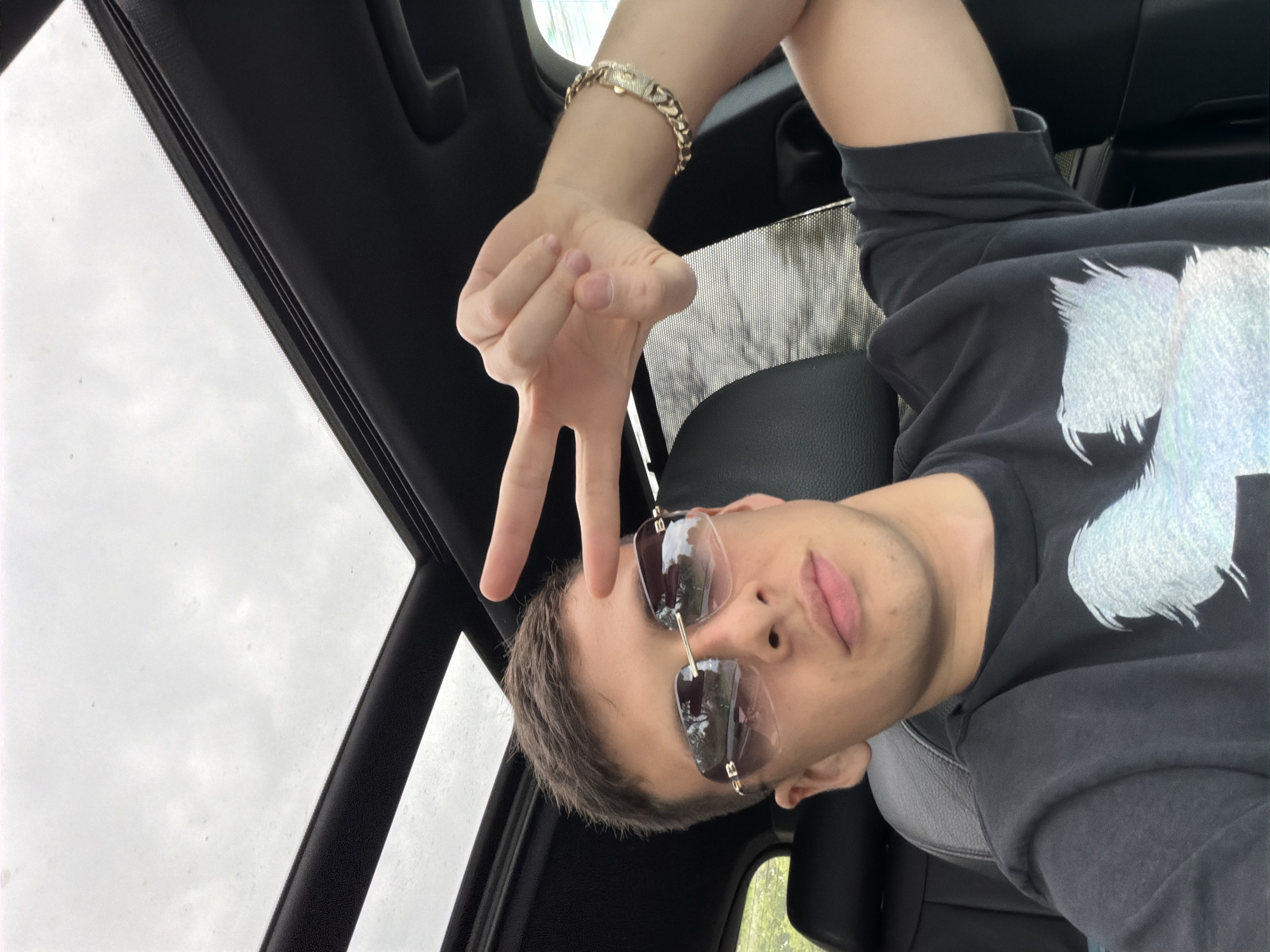 A young man is pictured from an overhead angle, sitting in what appears to be the driver's seat of a car. The image is oriented such that he appears to be looking upwards and slightly to his left. He has short, dark hair and is wearing sunglasses with dark, reflective lenses that obscure his eyes. His lips are slightly parted. He is wearing a dark-colored (likely black or dark gray) t-shirt or sweatshirt with a large, textured light-colored graphic design on the front, resembling a brushstroke or splatter pattern.
He holds up his right hand, extending his index and middle fingers in a "V" or peace sign, while his thumb, ring, and pinky fingers are curled. On his right wrist, he wears a chunky gold-colored chain bracelet with a decorative clasp.
The interior of the car is visible, featuring dark upholstery (possibly leather) on the seats and ceiling trim. Above him, a sunroof or panoramic roof allows a view of a bright, overcast sky with white and light gray clouds, indicating cloudy weather during the daytime. Reflections of the car's interior and the sky are visible in the lenses of his sunglasses. No visible text is present in the image.
